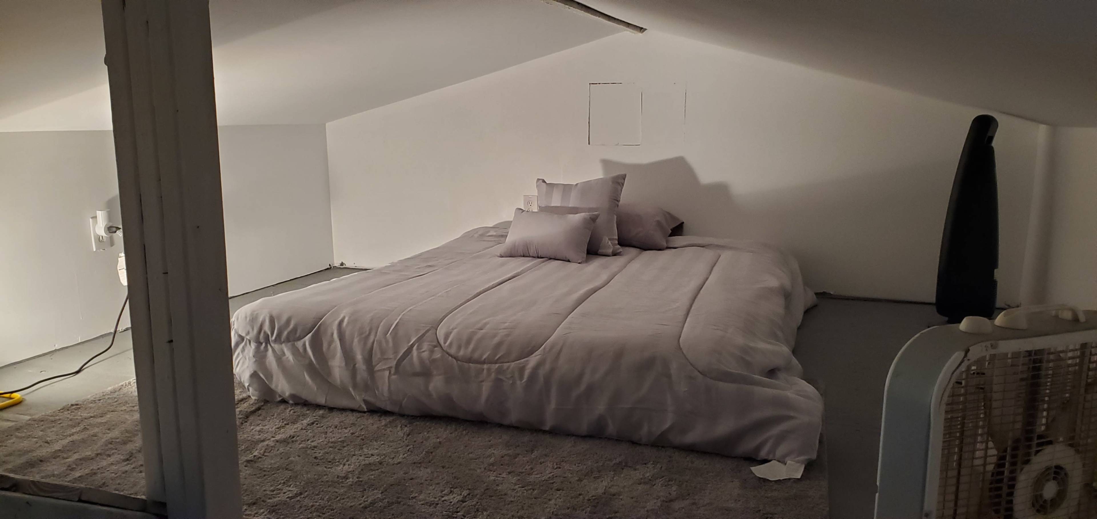 The image shows a sparsely furnished attic bedroom featuring a large bed covered with a light gray comforter and several pillows against a white wall.