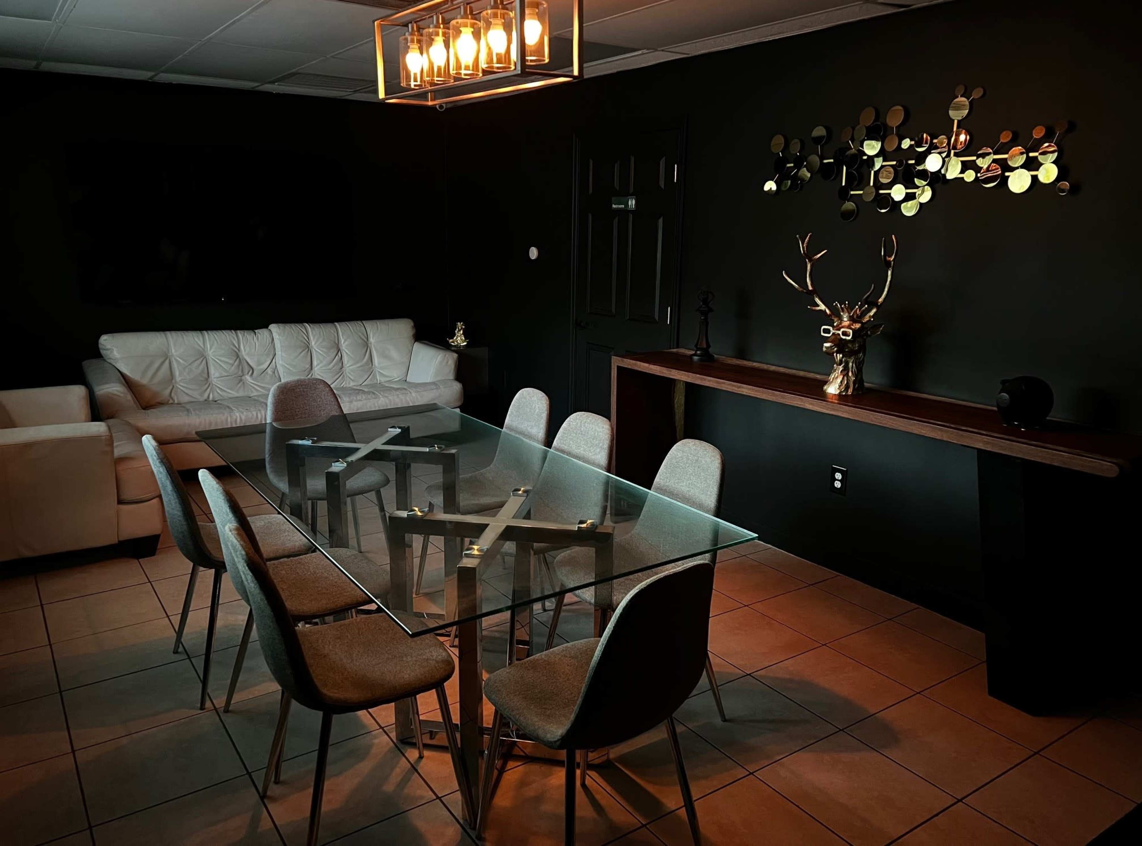 A modern dining area features a glass table surrounded by six chairs, with a white couch and decorative wall art in a dark room.