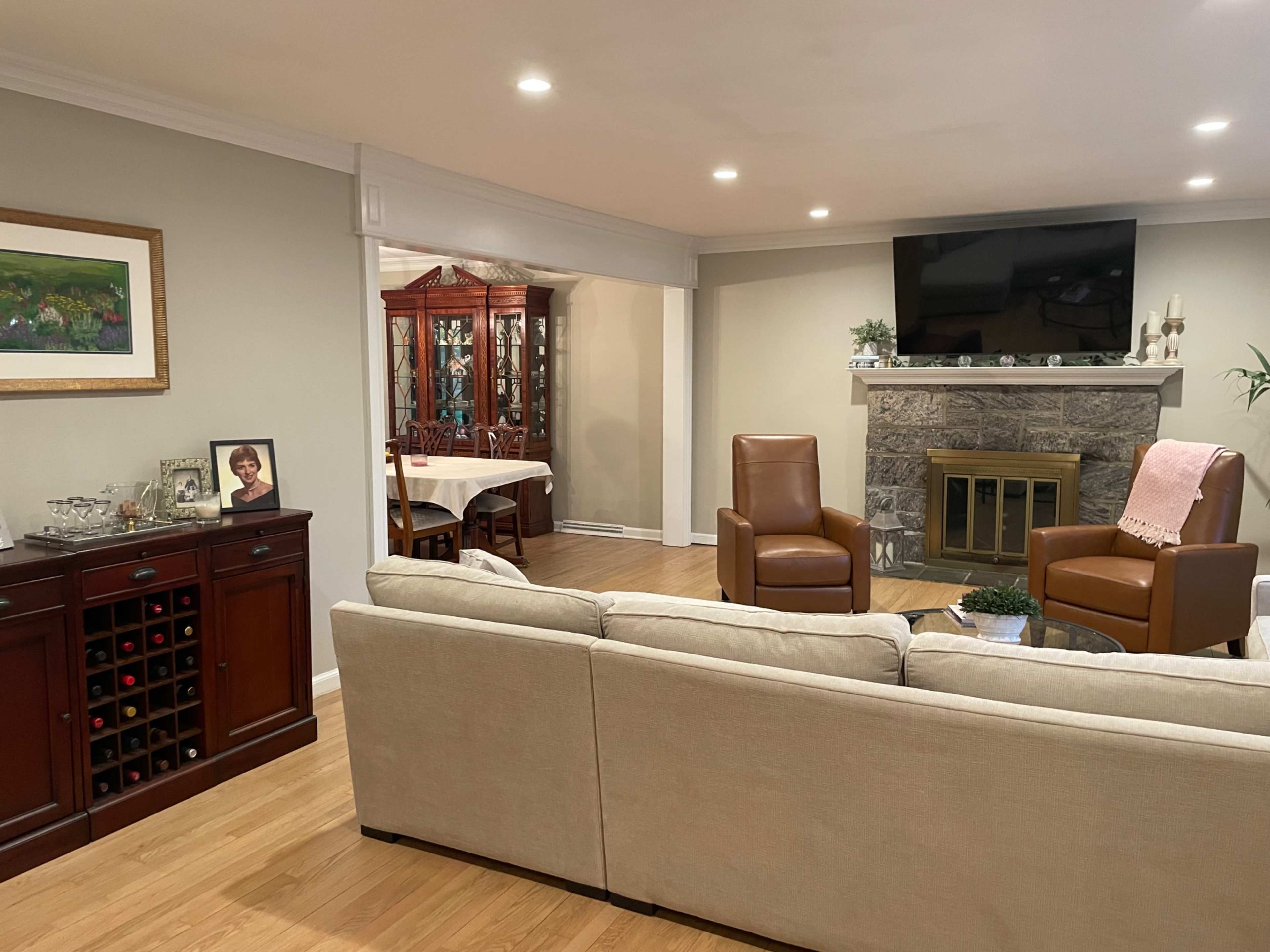 A cozy living room features a beige sectional sofa, two brown chairs, a stone fireplace, and a wall-mounted television, with a dining area visible in the background.