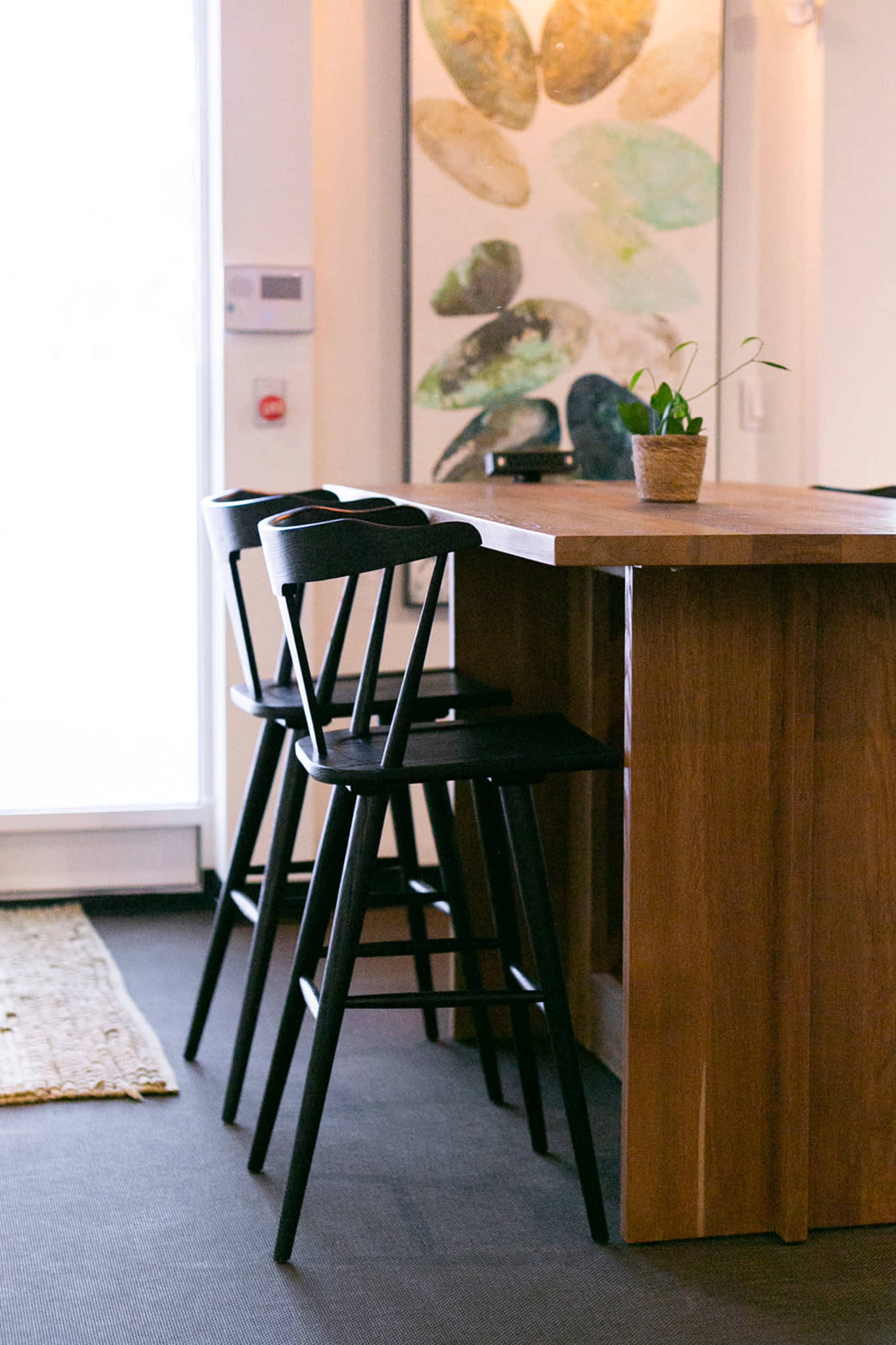 The image features a wooden kitchen island with three black chairs and a potted plant on top, set against a wall adorned with abstract art.