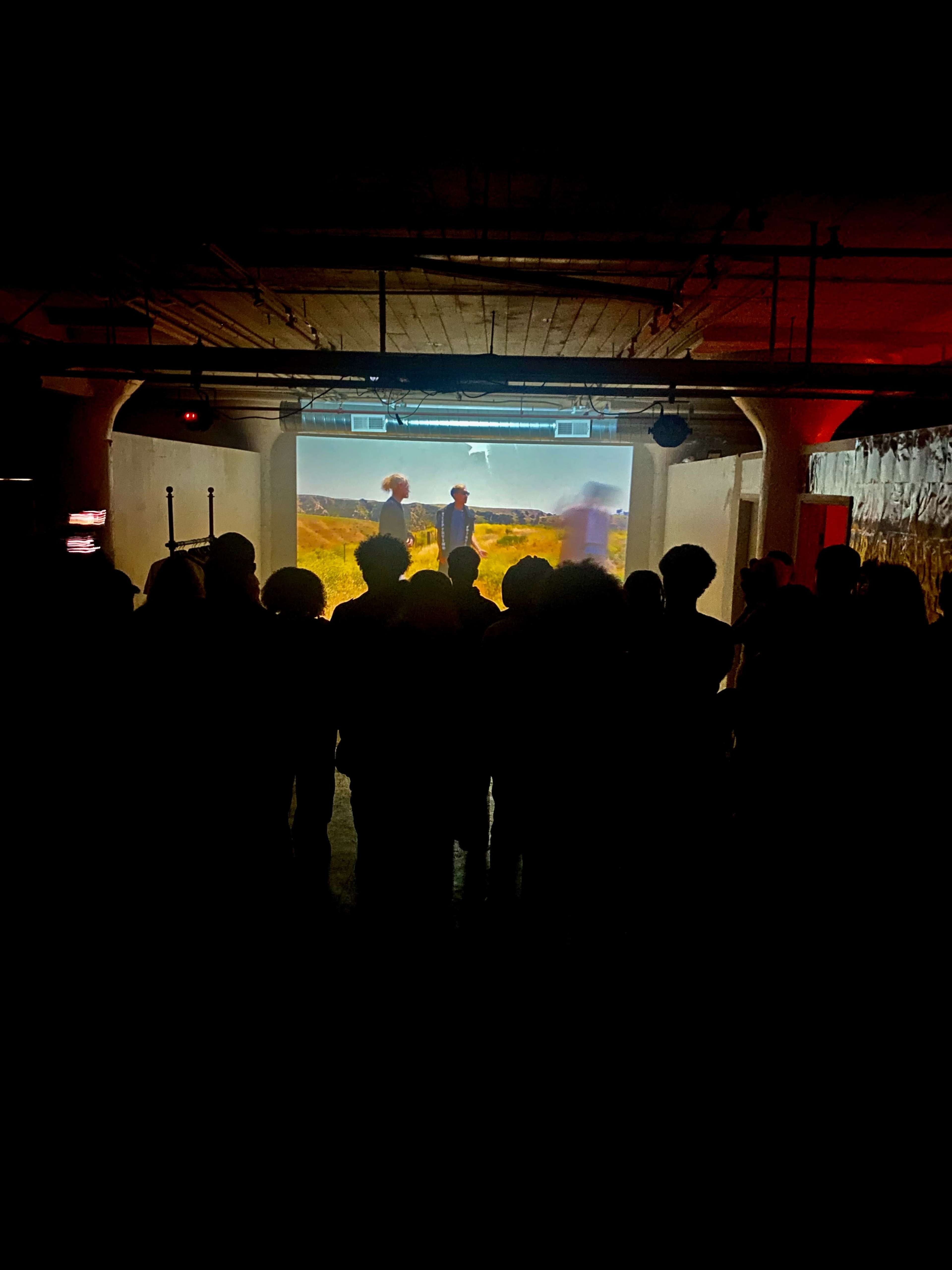 A group of people watches a projection on a screen in a dimly lit room.