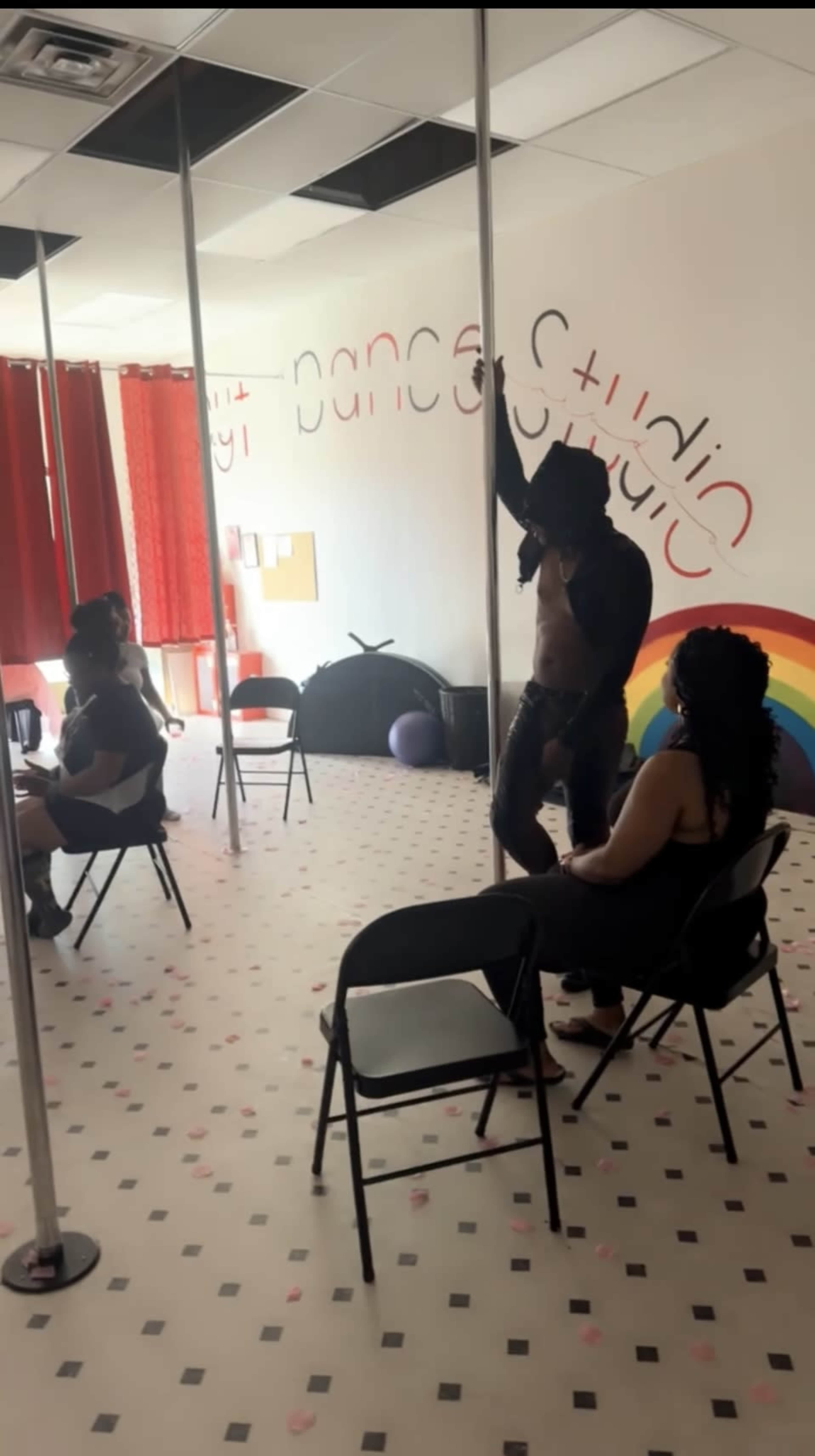 A person performs a pole dance in a studio with three seated spectators and colorful decor on the walls.