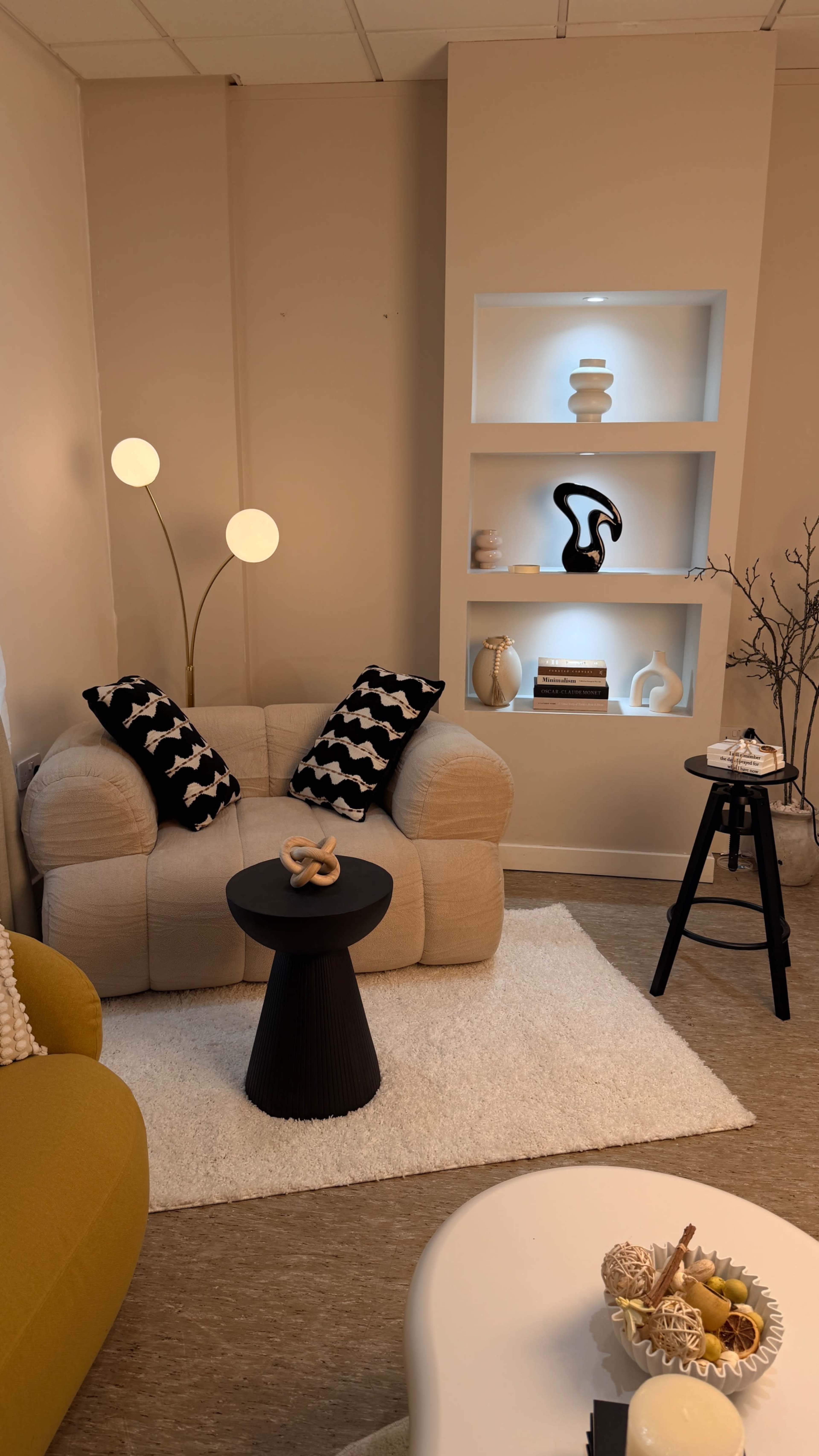 The image shows a cozy living room corner featuring a beige couch with black and white throw pillows, a small black side table, and decorative shelves with various items.