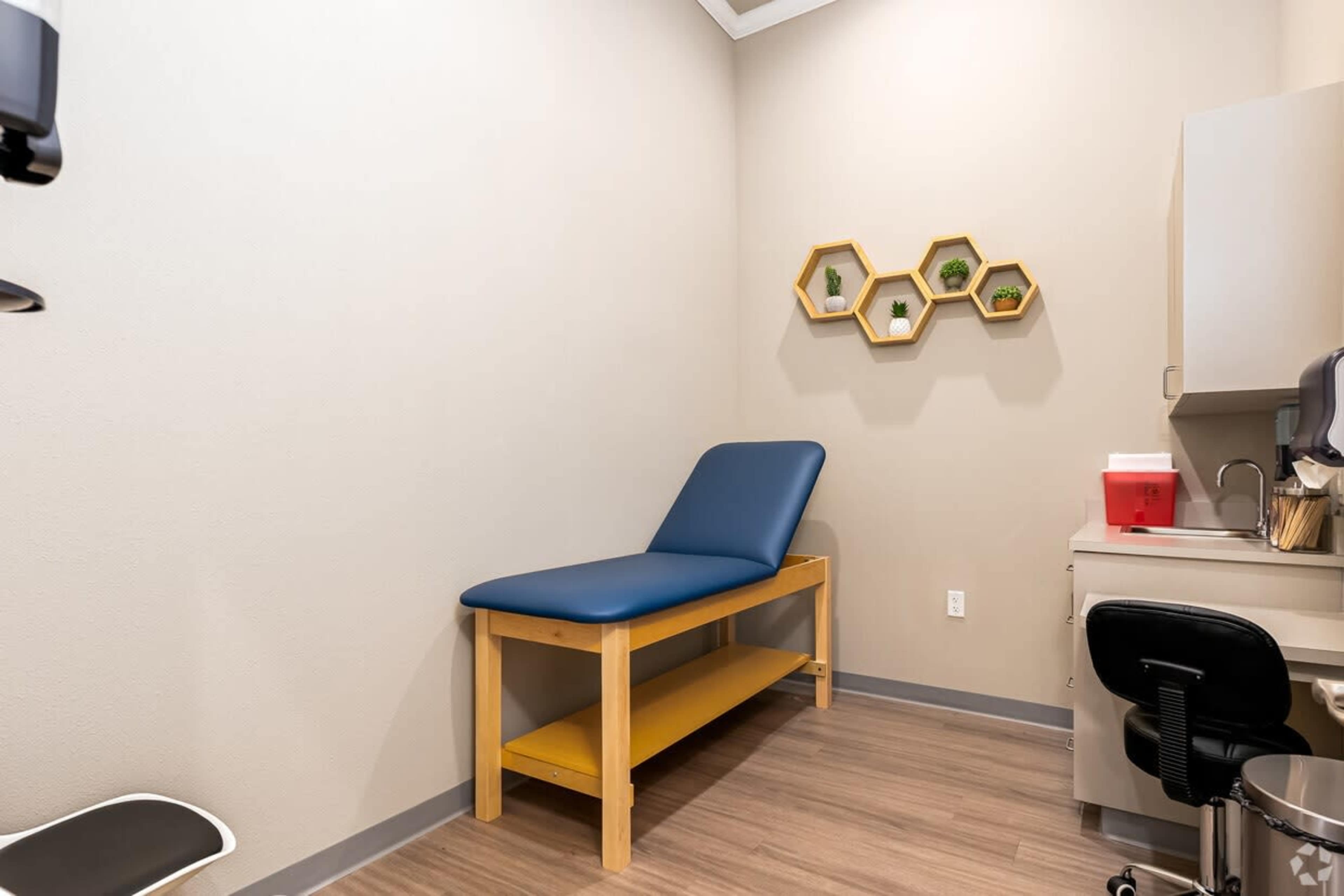 A medical examination room with a blue exam table, a small side shelf, and a chair next to a desk.