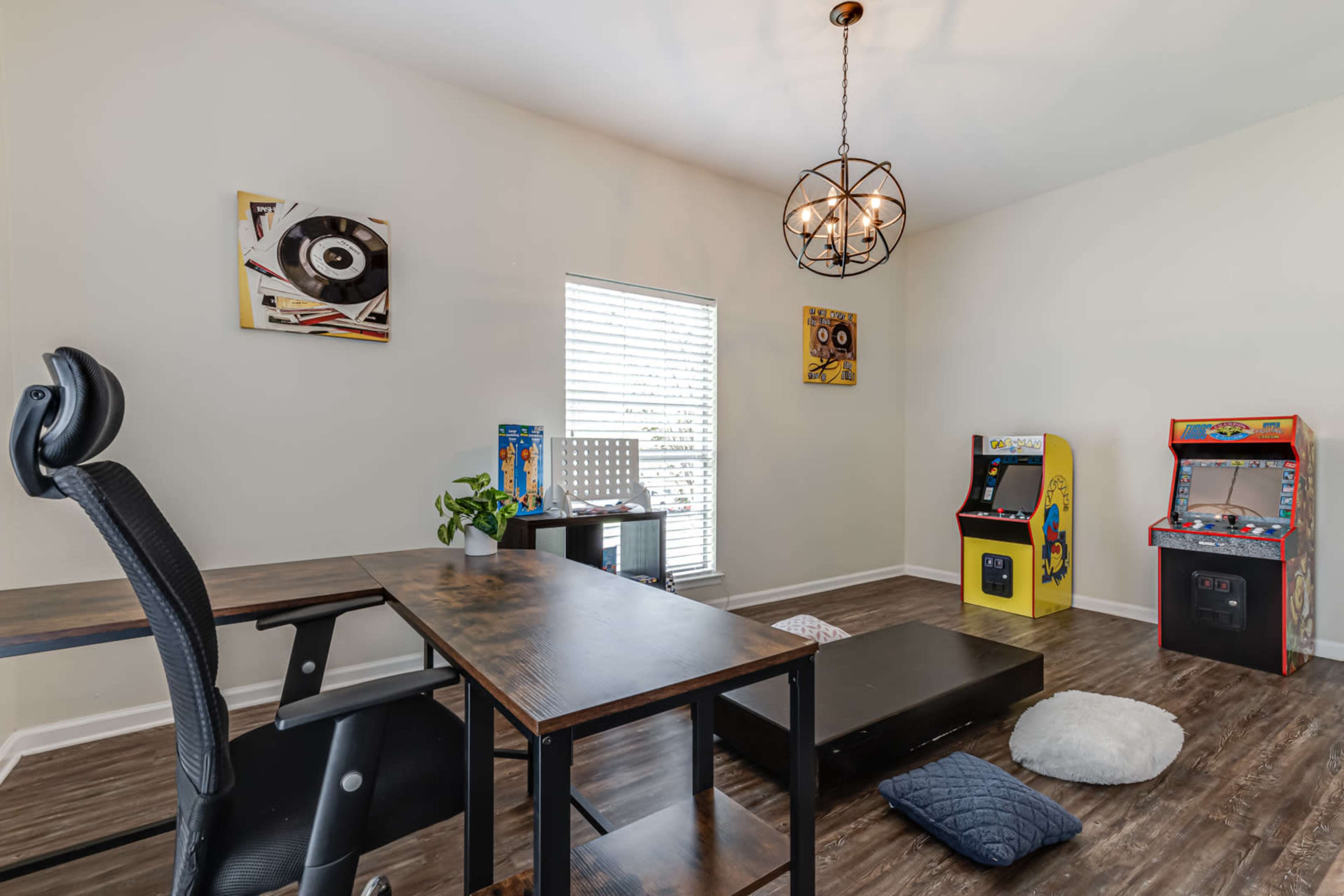A home office features a large desk, an office chair, a plant, and two vintage arcade machines against a wall with framed artwork.