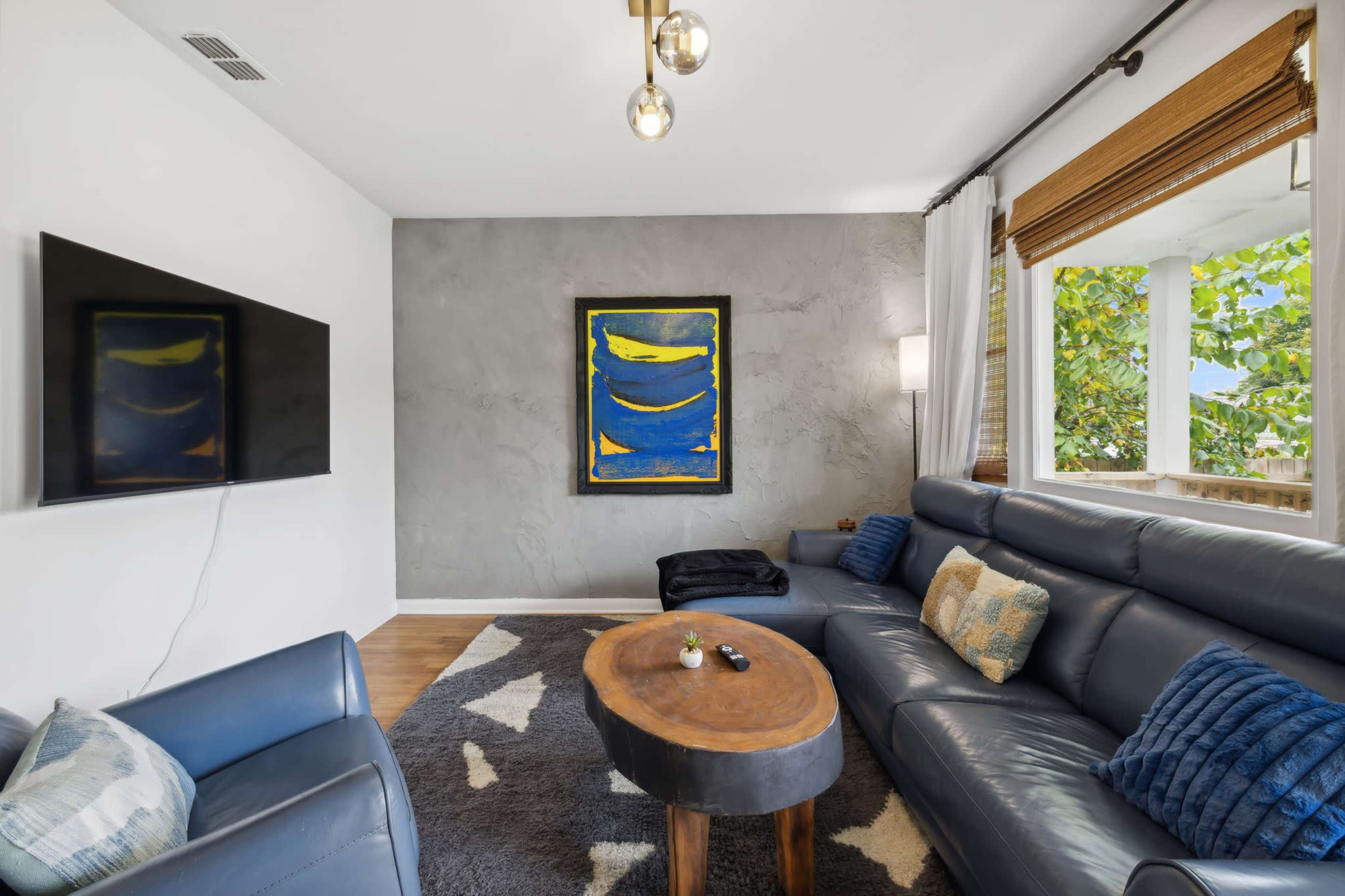A modern living room features a dark blue leather sofa, a wooden coffee table, and a large abstract painting on the wall, with a flat-screen TV mounted nearby.