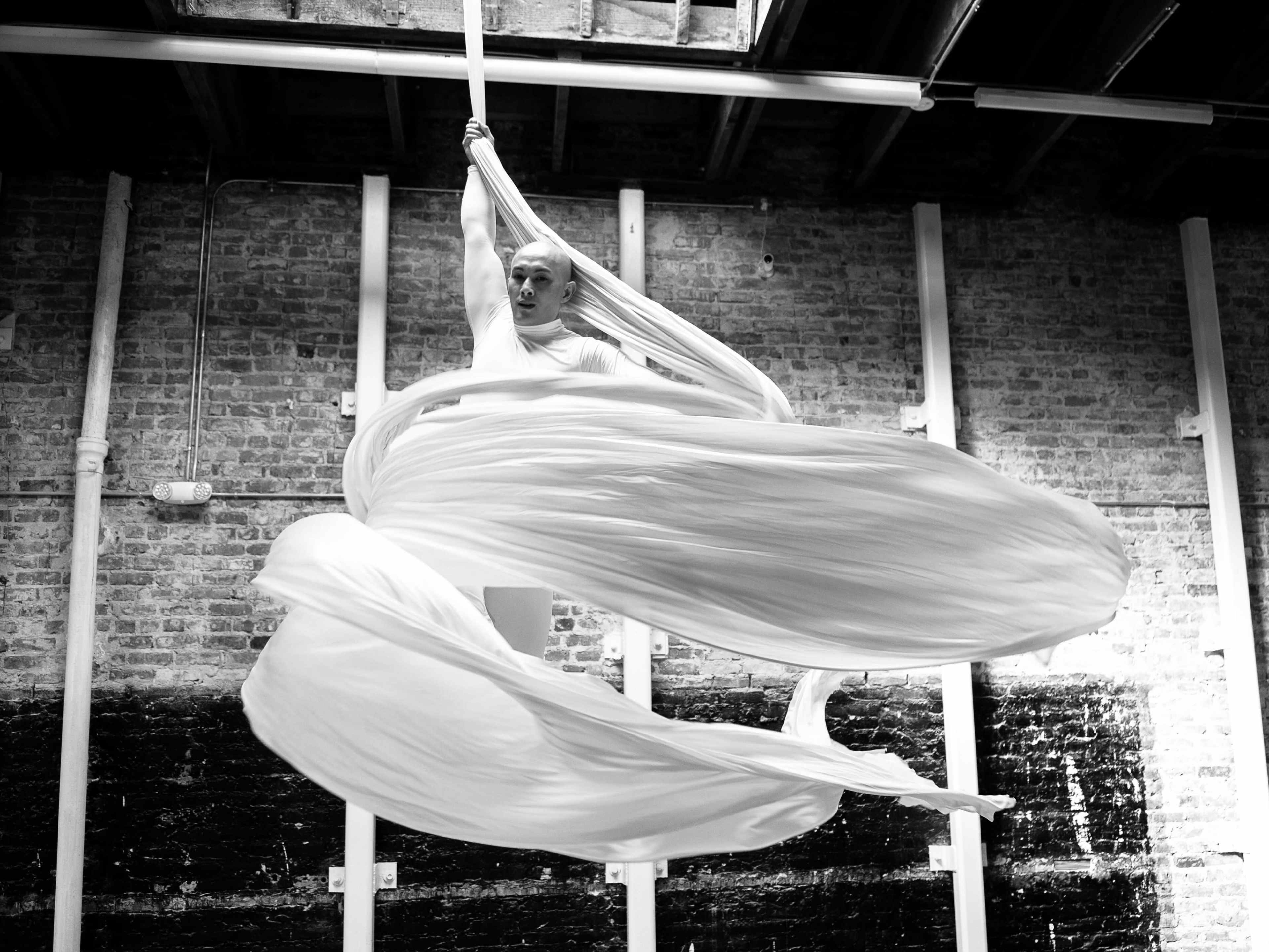 A performer spins gracefully in midair while suspended from silk fabric in a brick-walled studio.
