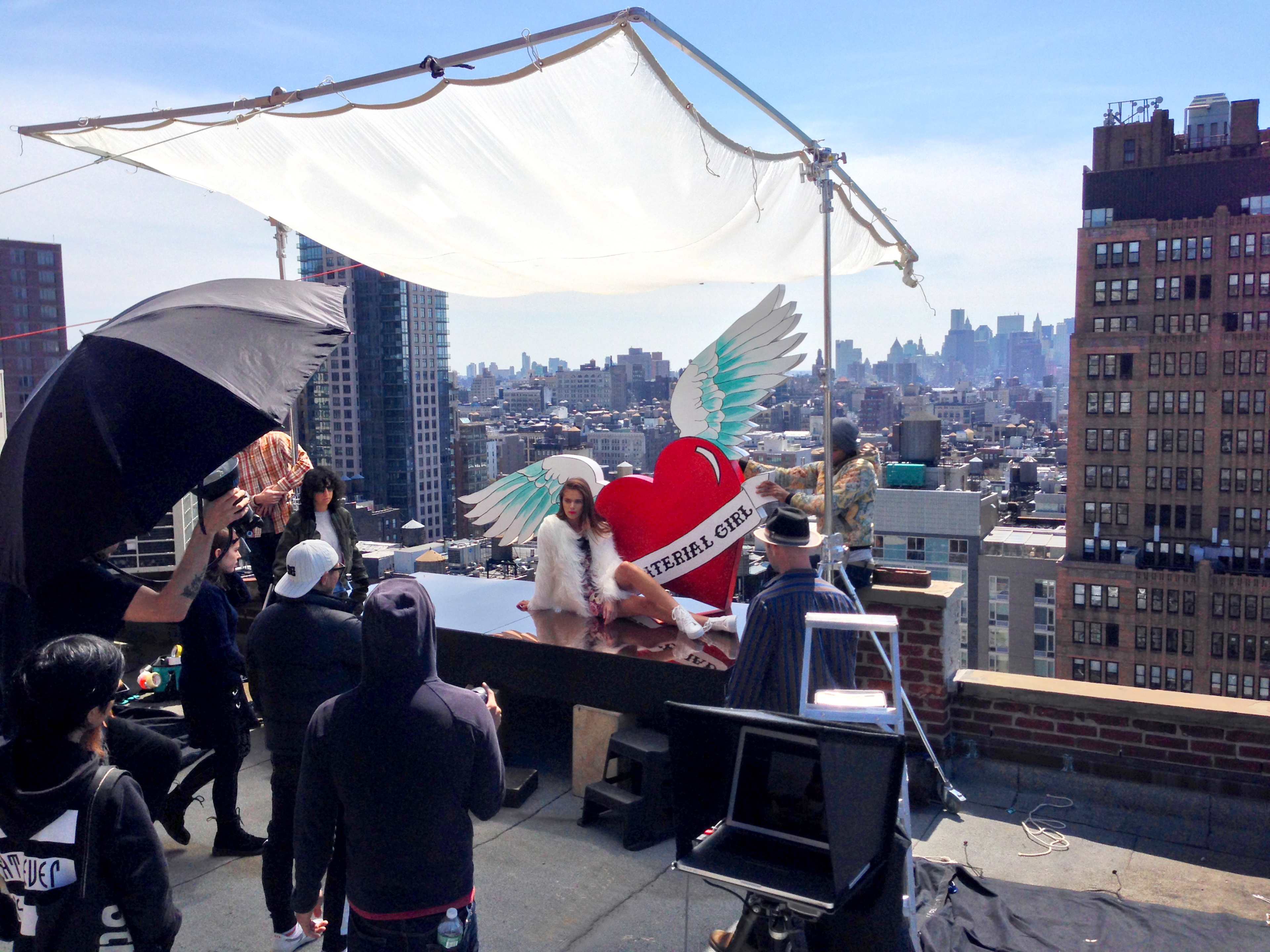 A photoshoot is taking place on a rooftop with city skyline views, featuring a model with angel wings and a large heart backdrop.