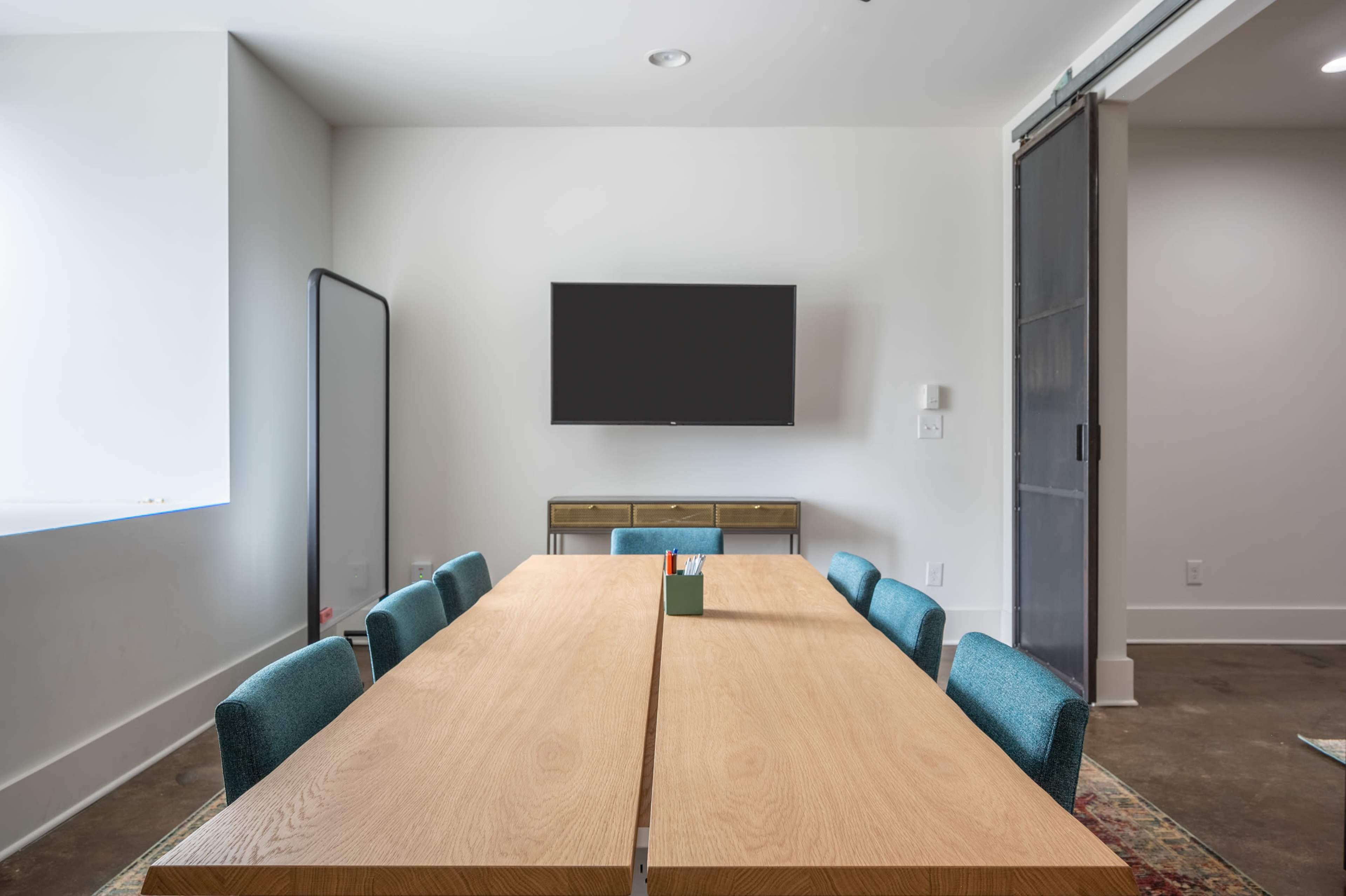 A modern meeting room features a wooden table with eight teal chairs, a wall-mounted television, and a large mirror.