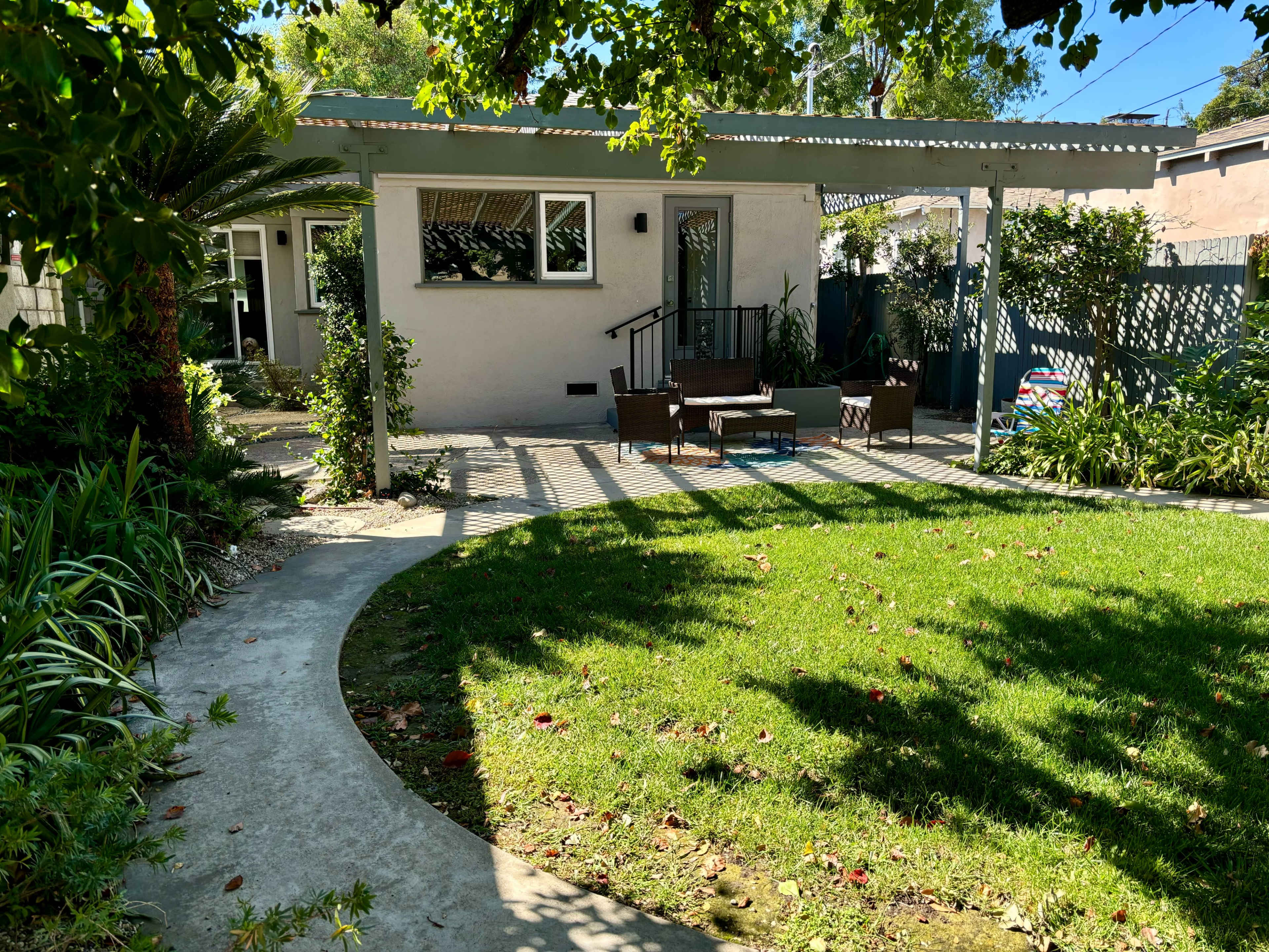 The image shows a small, well-maintained backyard with a green lawn, a concrete pathway, and a covered porch area adjacent to a light-colored house.