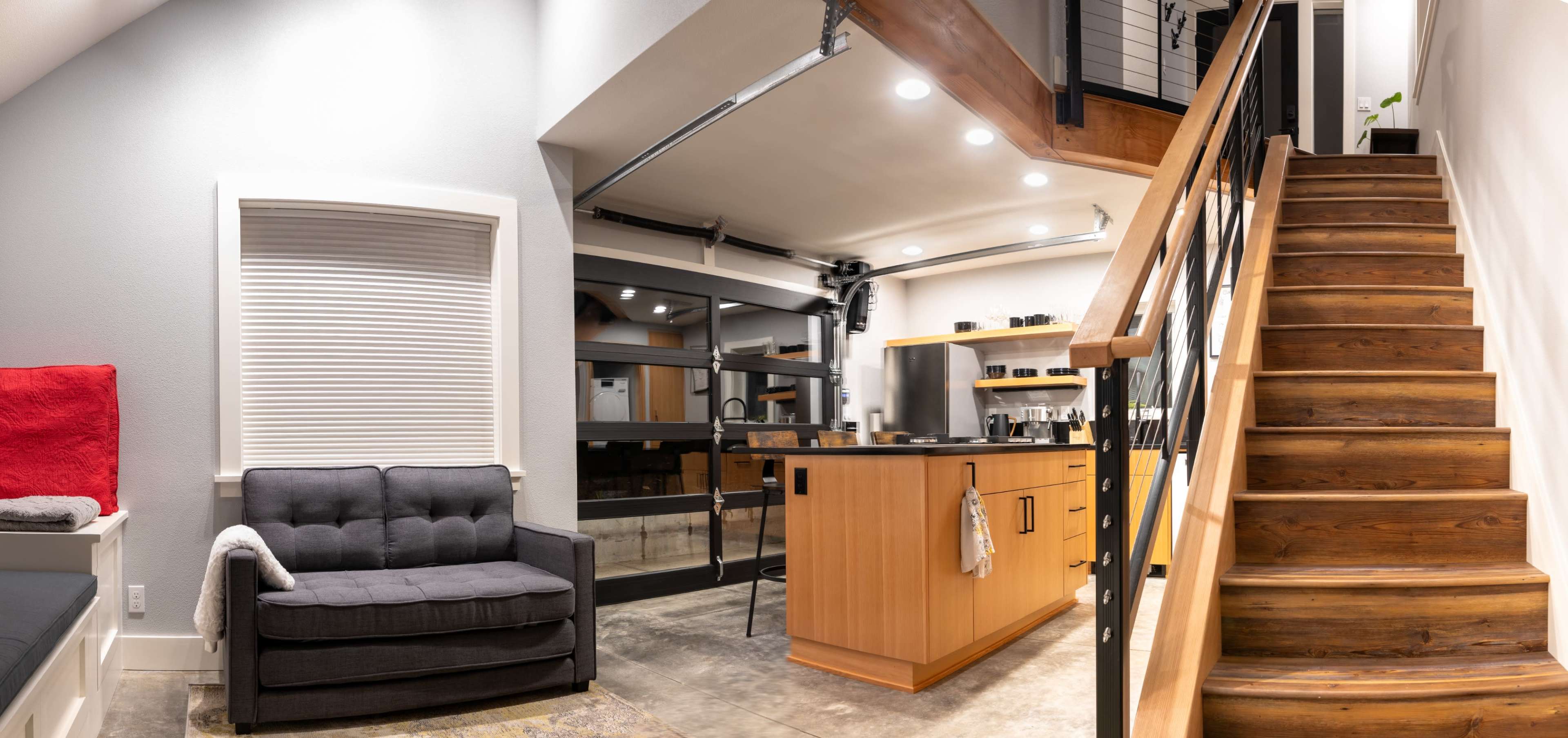The image shows a modern interior with a staircase leading to an upper level, a gray couch on the left, and a kitchen area with wooden cabinetry and stainless steel appliances.