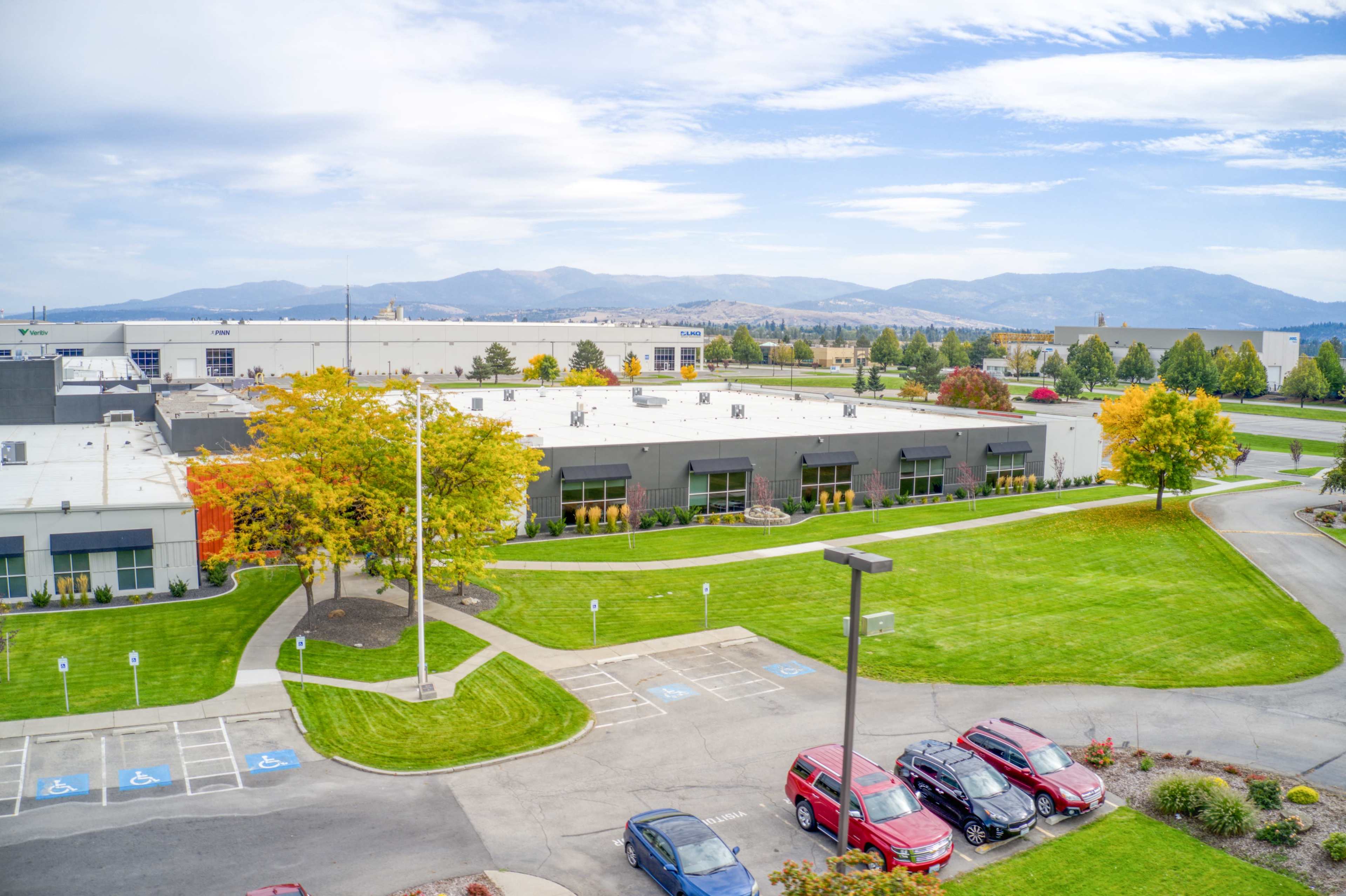 A modern commercial building is surrounded by well-maintained lawns and parking spaces, with mountains visible in the background.