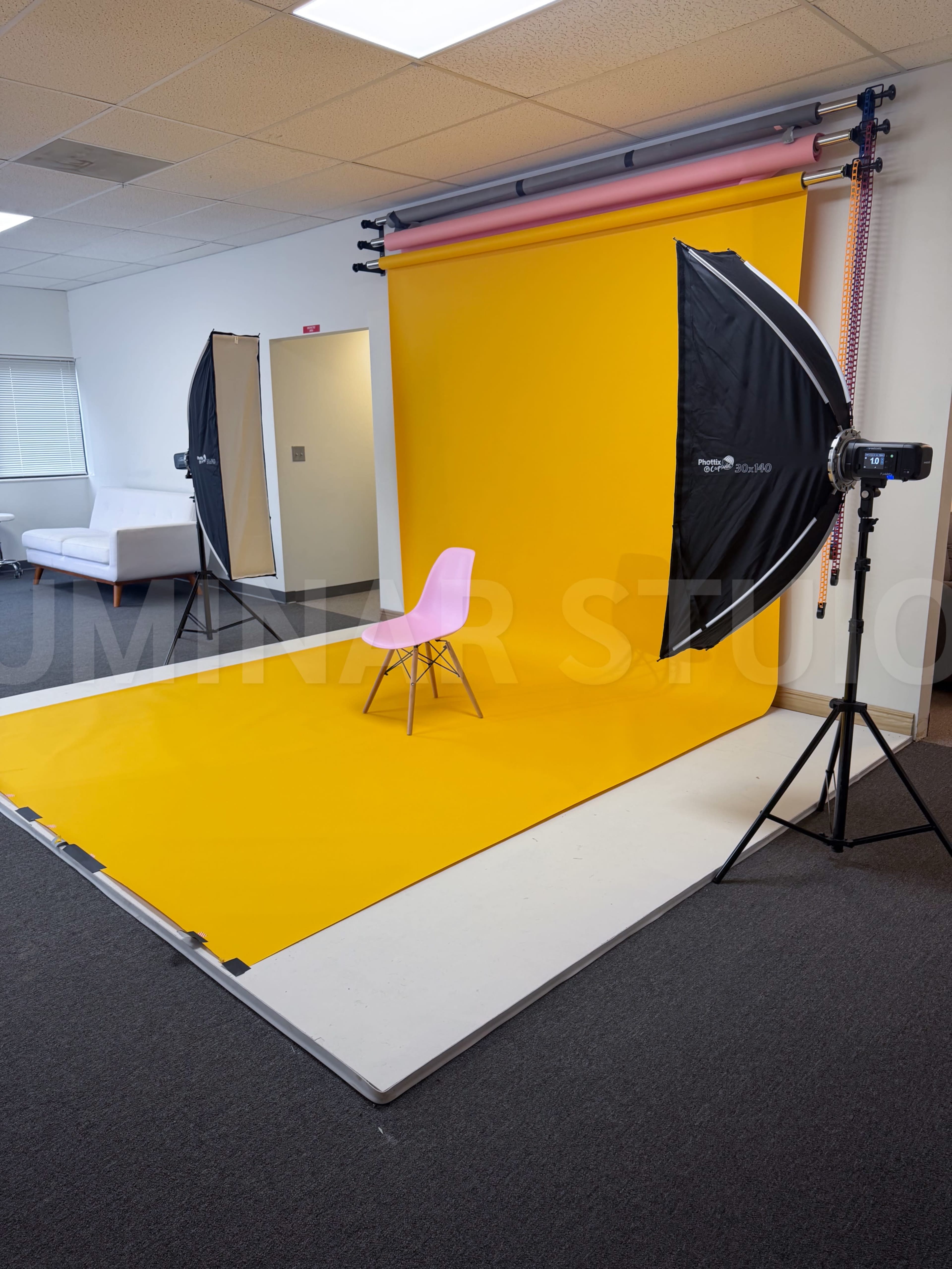 The image shows a photography studio setup with a bright yellow backdrop, a single pink chair in the center, and lighting equipment positioned on either side.