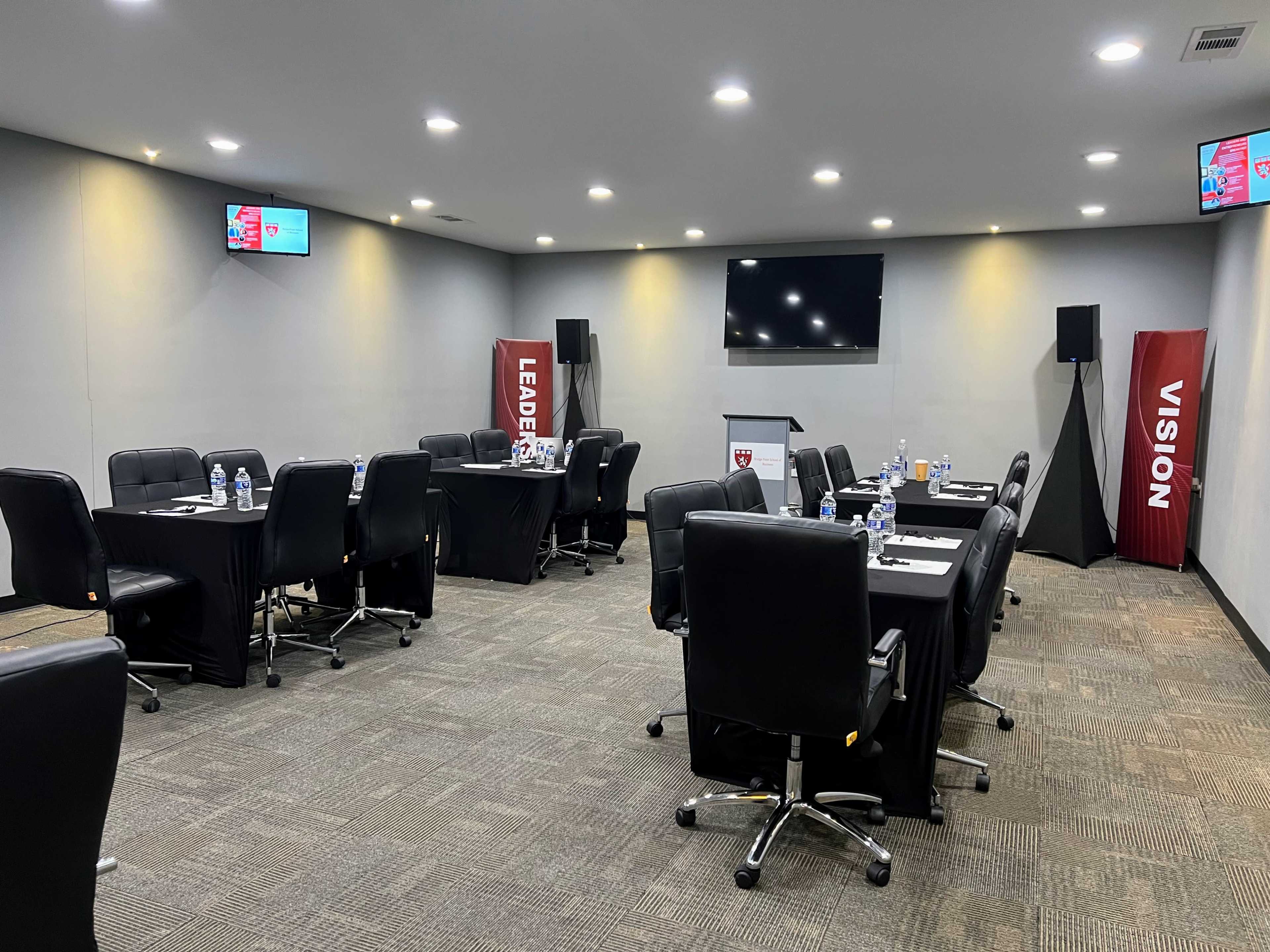 The image shows a conference room set up with several tables and chairs, a large screen, and banners displaying the words "LEADERSHIP" and "VISION."