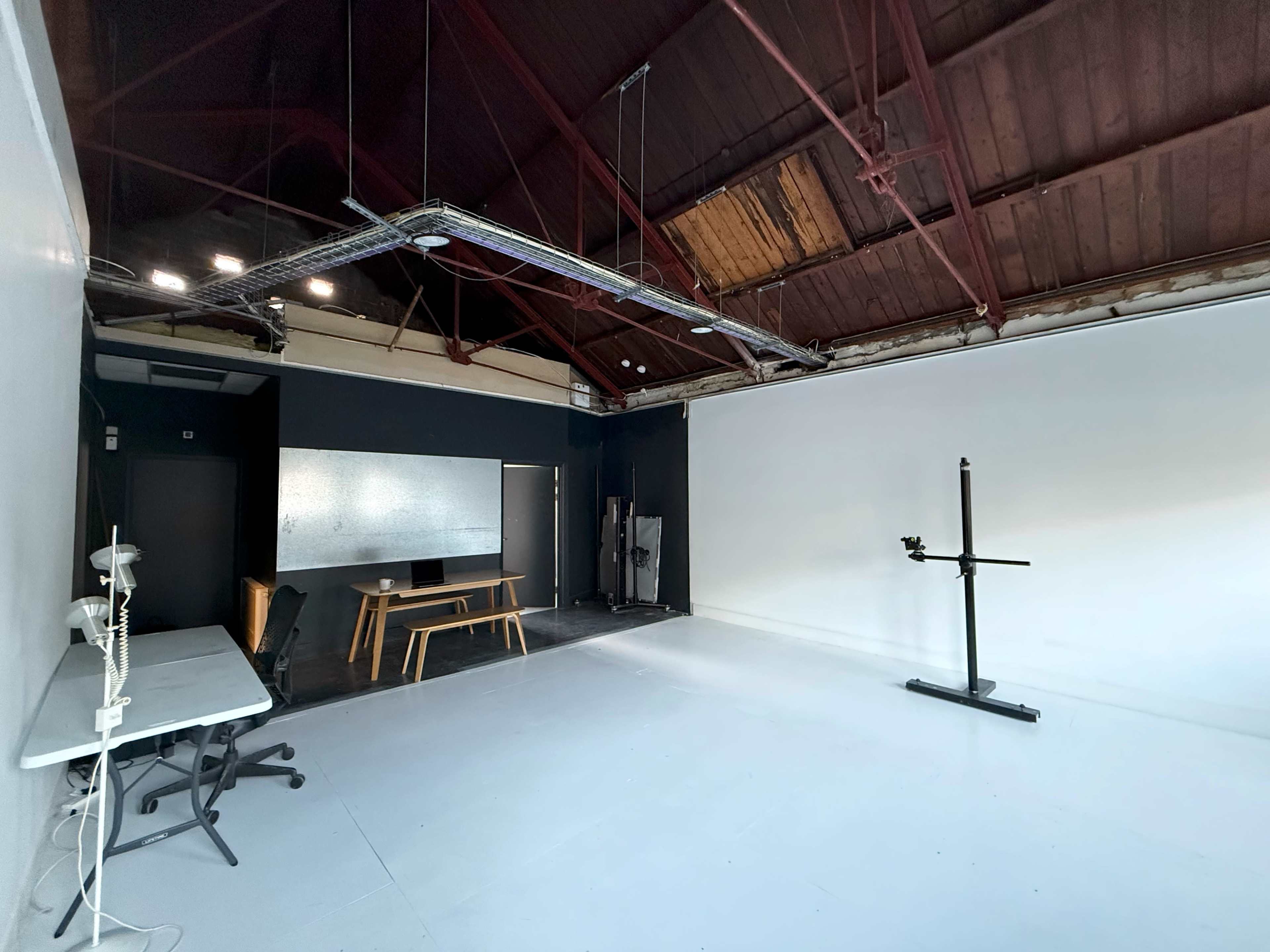 The image shows an empty studio space with a white floor, a black wall, a large white backdrop, a table with chairs, and a lighting setup on the ceiling.