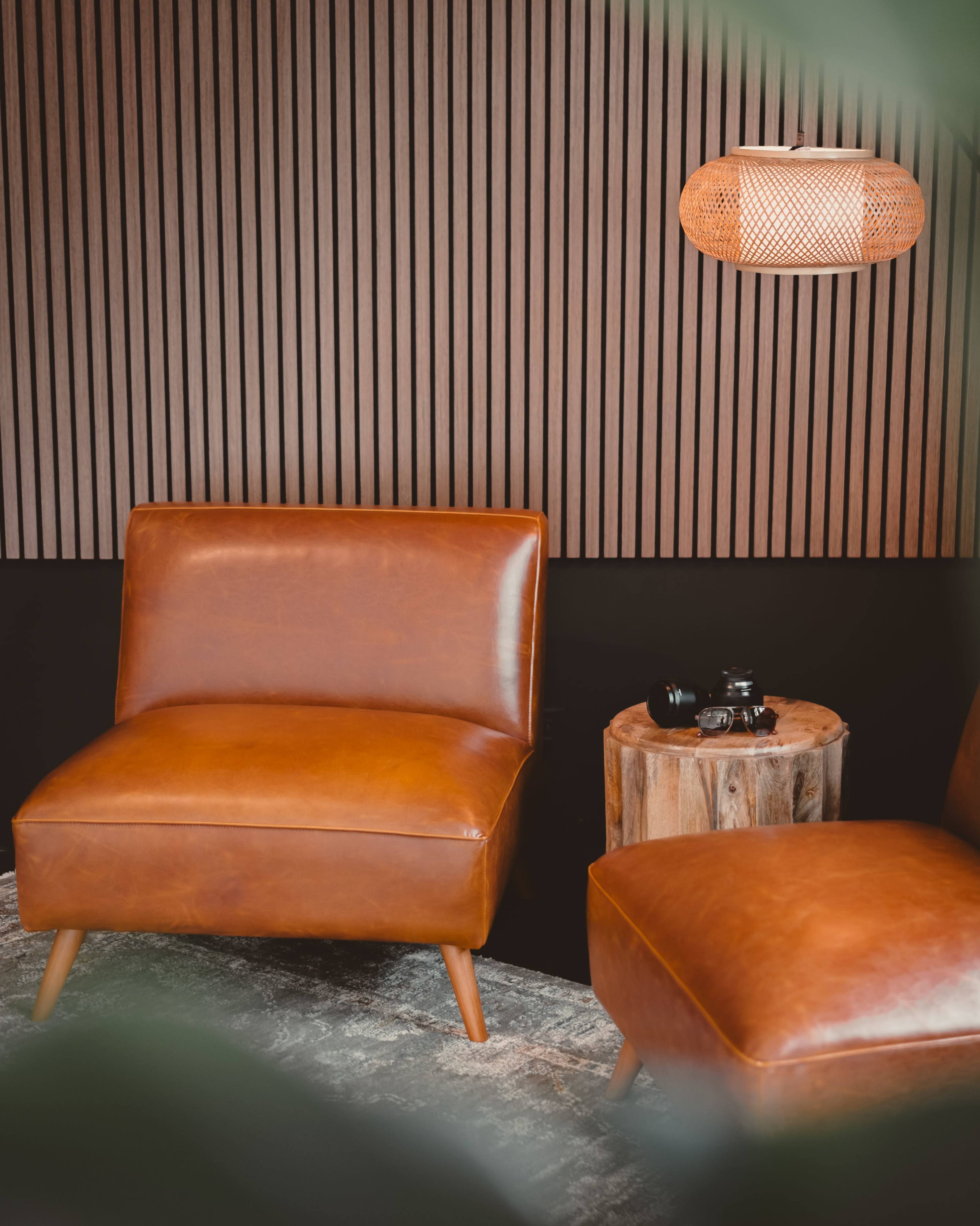 A cozy seating area with two brown leather chairs, a cylindrical wooden table, and a woven light fixture against a striped wooden wall.