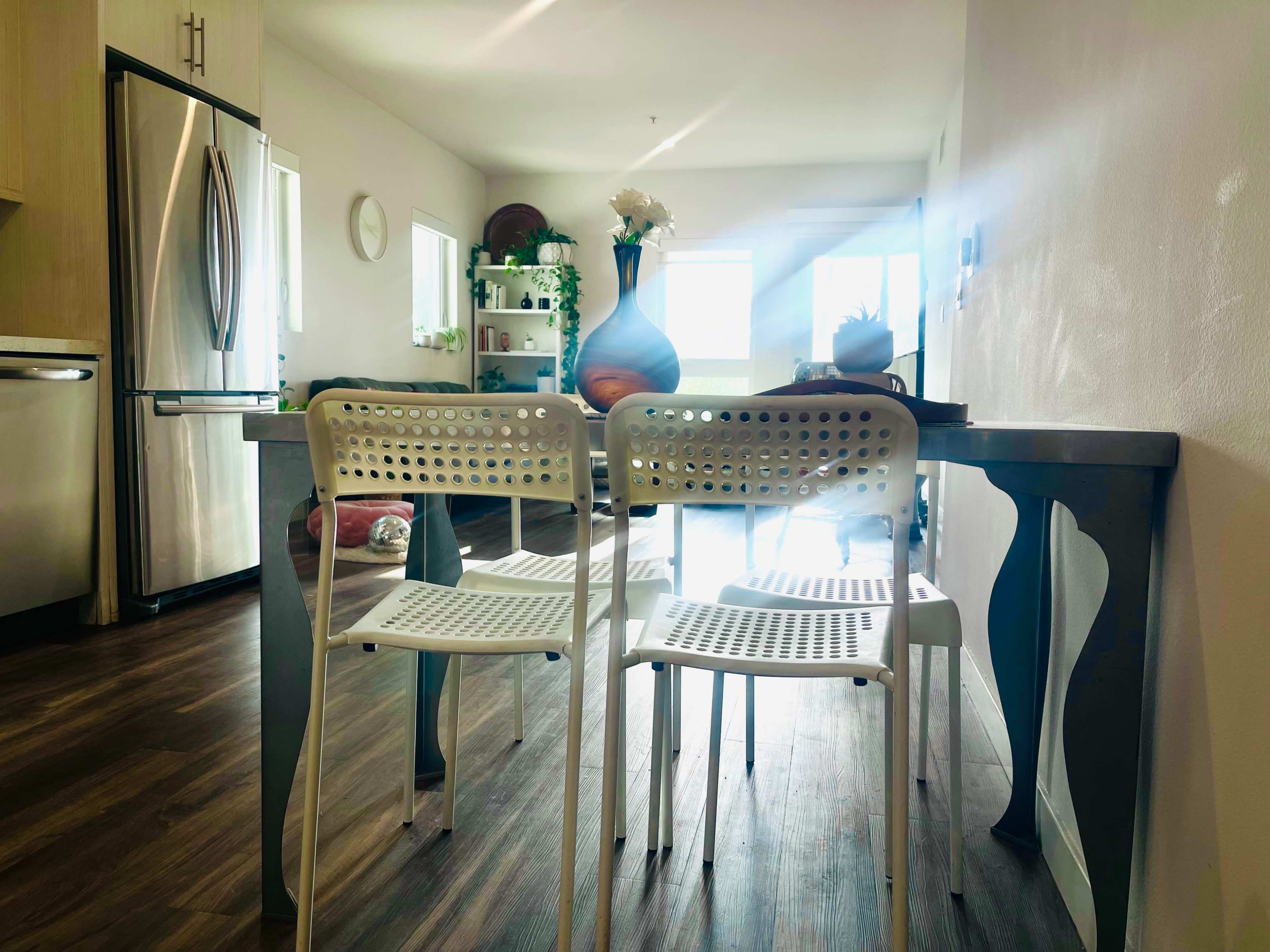 A modern kitchen scene features a table with two white chairs, illuminated by natural light coming through a window, with decorative items on display in the background.