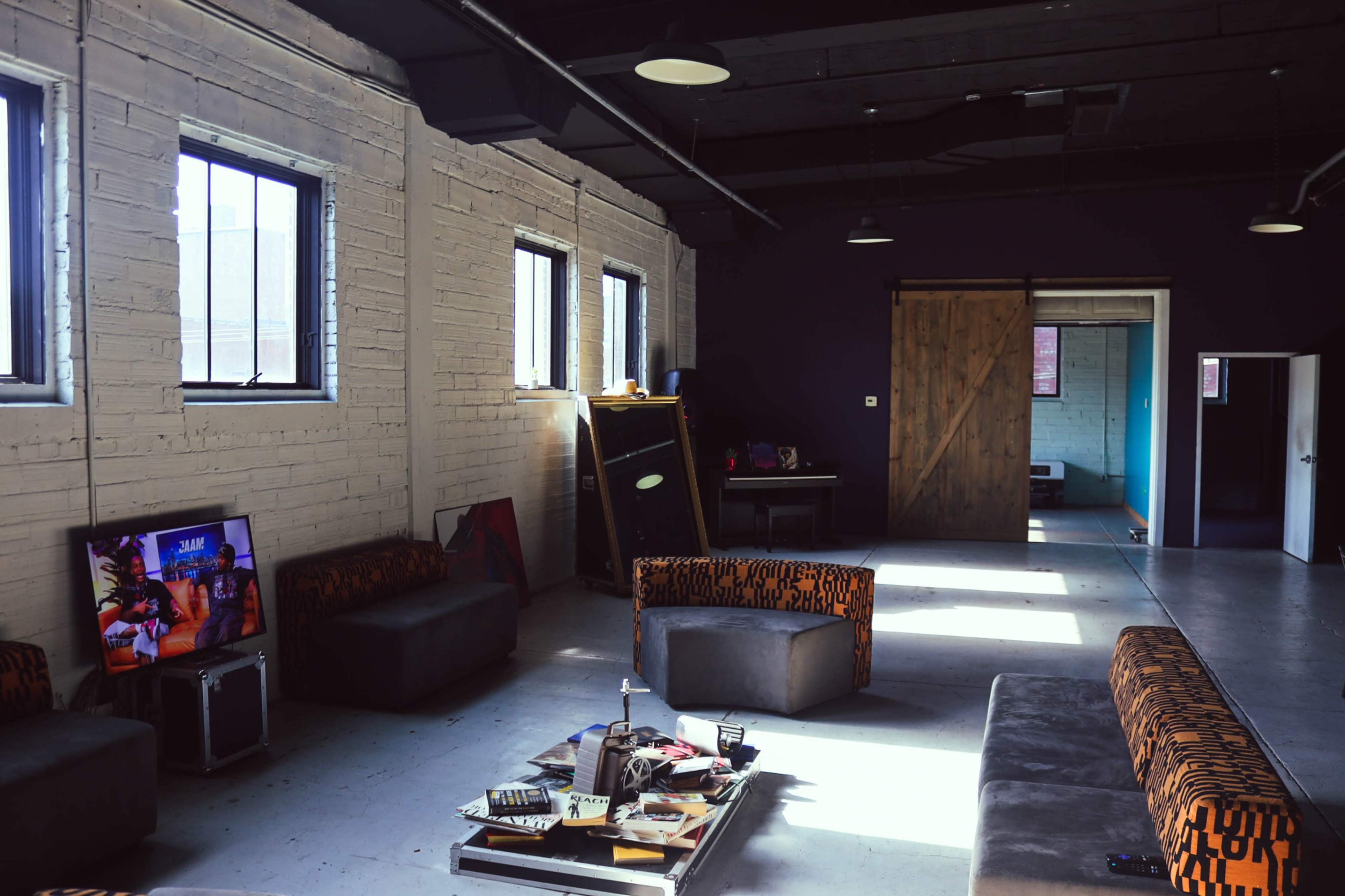 The interior of a spacious room features two large windows, a collection of low sofas with orange and black patterns, a coffee table with scattered items, and an open doorway leading to another area.