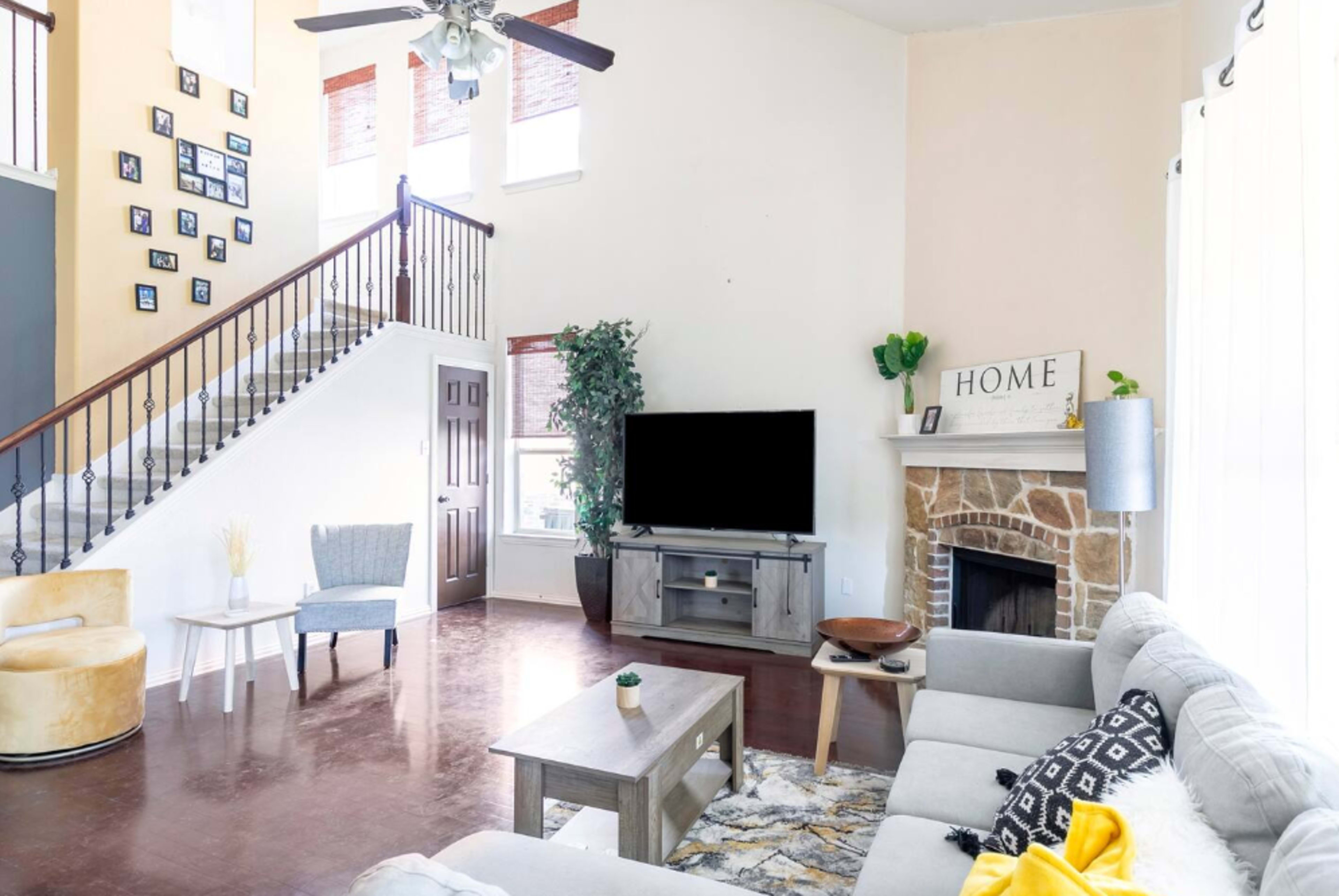 A spacious living room features a large television, a stone fireplace, and a staircase adorned with family photos.