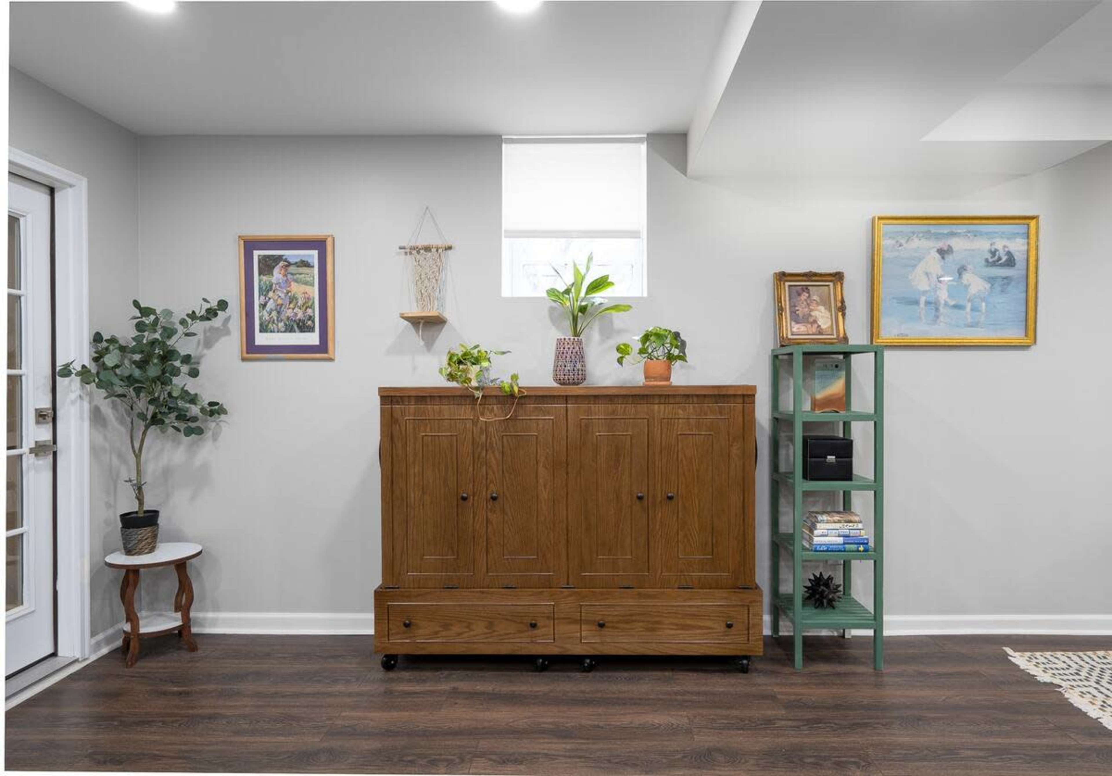 A wooden cabinet with drawers is placed against a wall adorned with a painting and a framed picture, while a plant and a small shelf with more decor items are nearby.