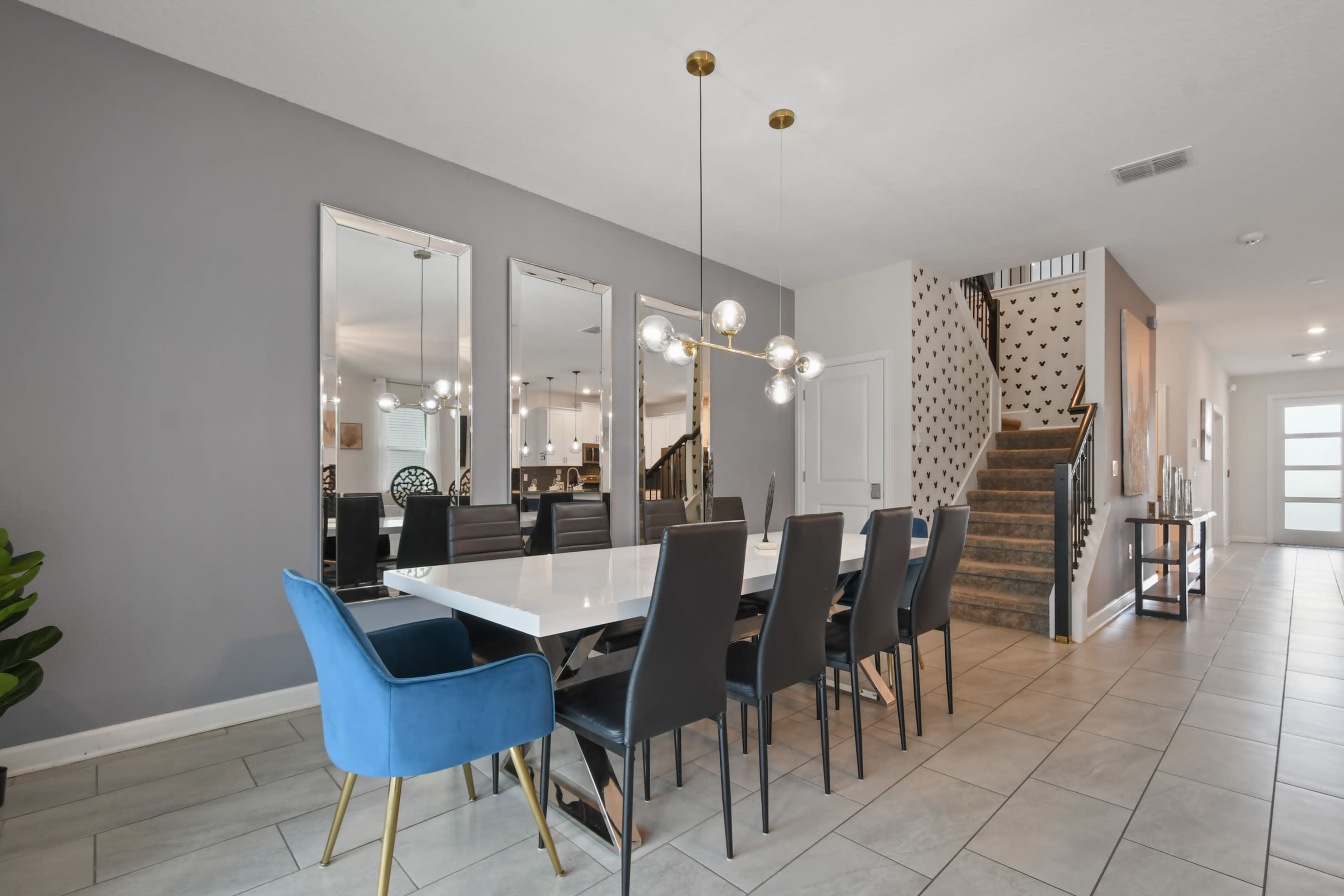 A modern dining area featuring a long white table surrounded by black chairs, with a blue accent chair, large mirrors on the wall, and a staircase in the background.