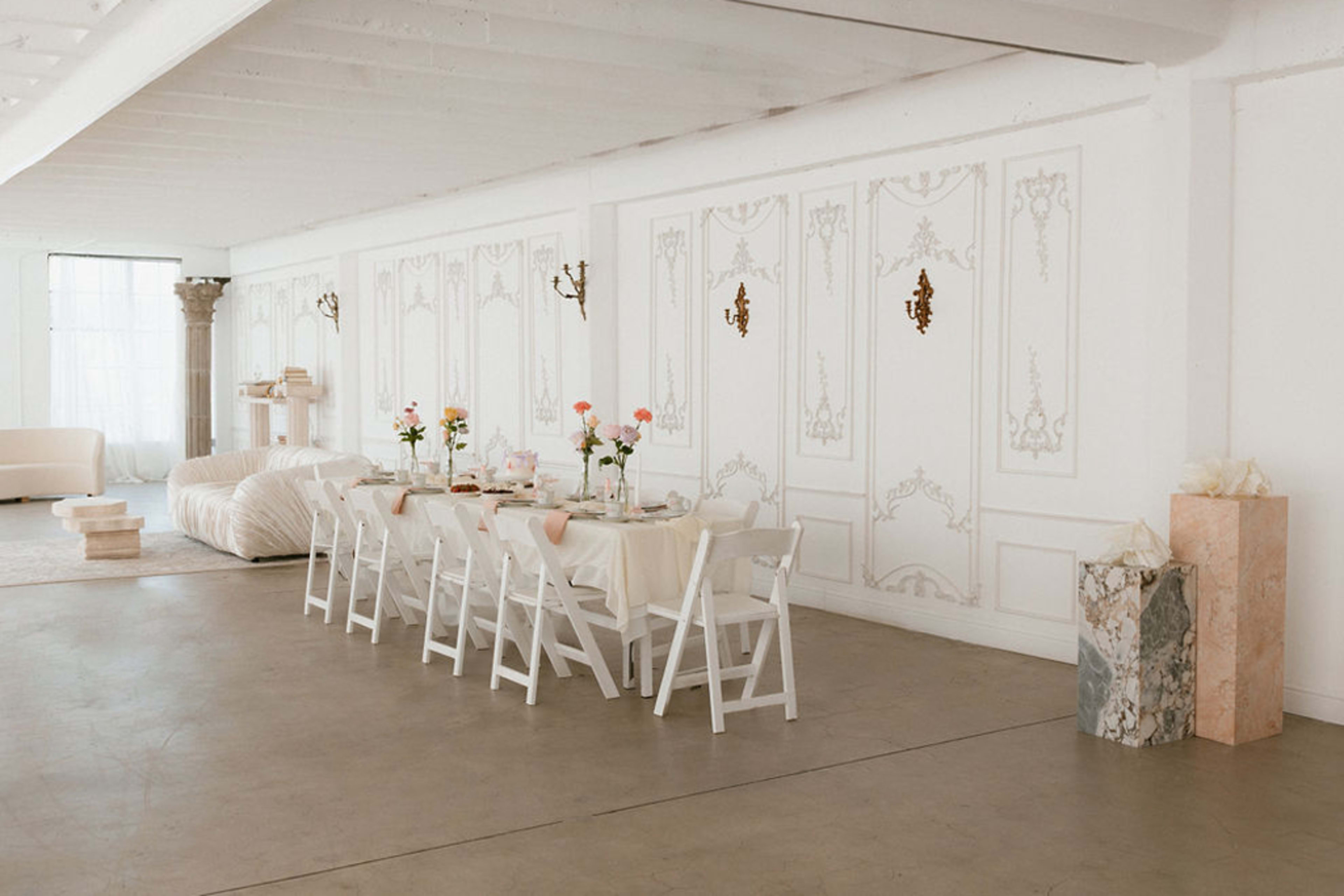 A long, elegantly set dining table with white chairs is positioned in a bright, minimalistic room featuring decorative wall moldings and soft natural light.