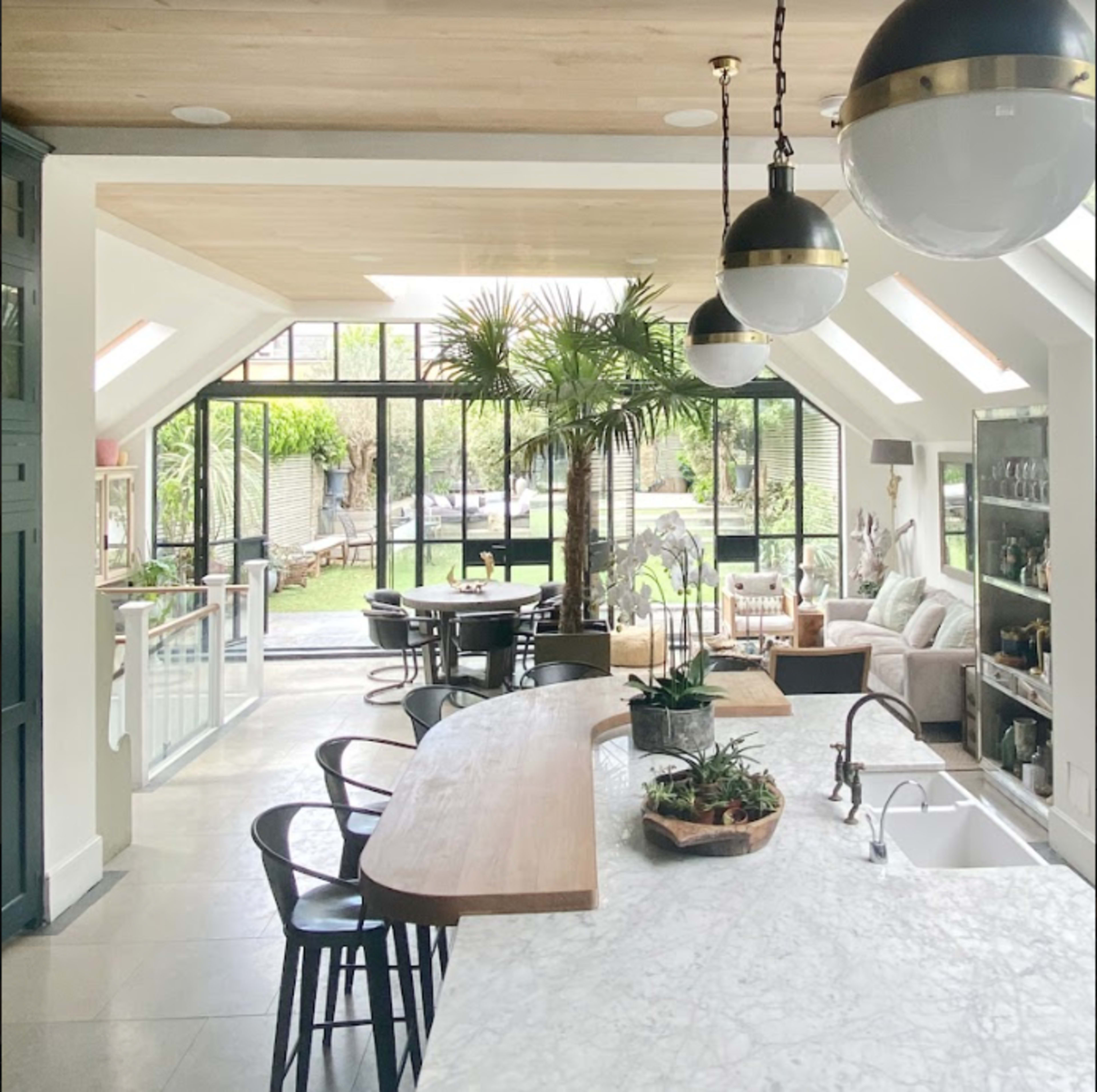 A spacious, modern kitchen and living area with large windows that open to a green outdoor space, featuring a central island with a marble countertop and a dining table.