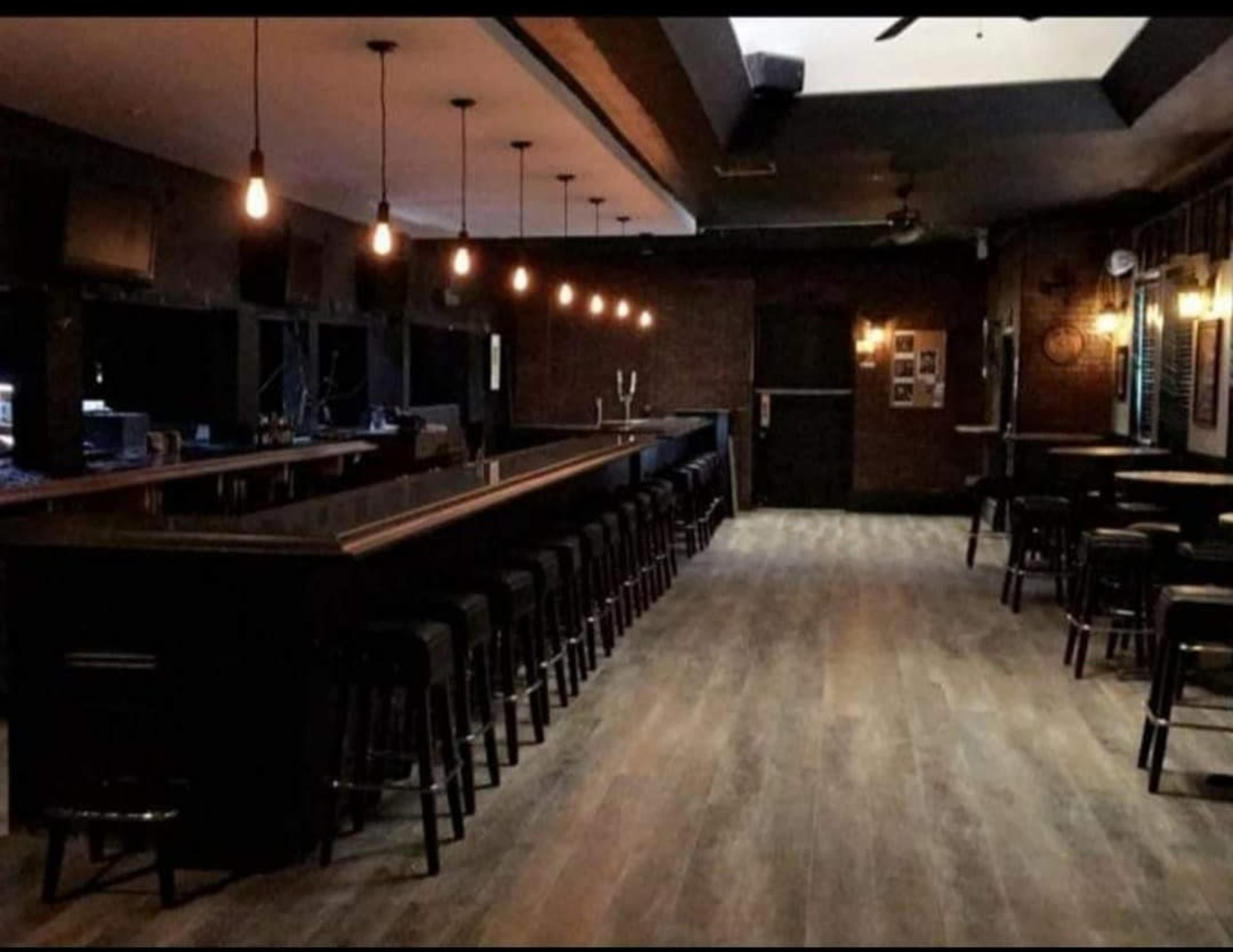 A dimly lit bar area with a long wooden counter and rows of black stools along the front, set against a backdrop of exposed brick walls and wooden flooring.