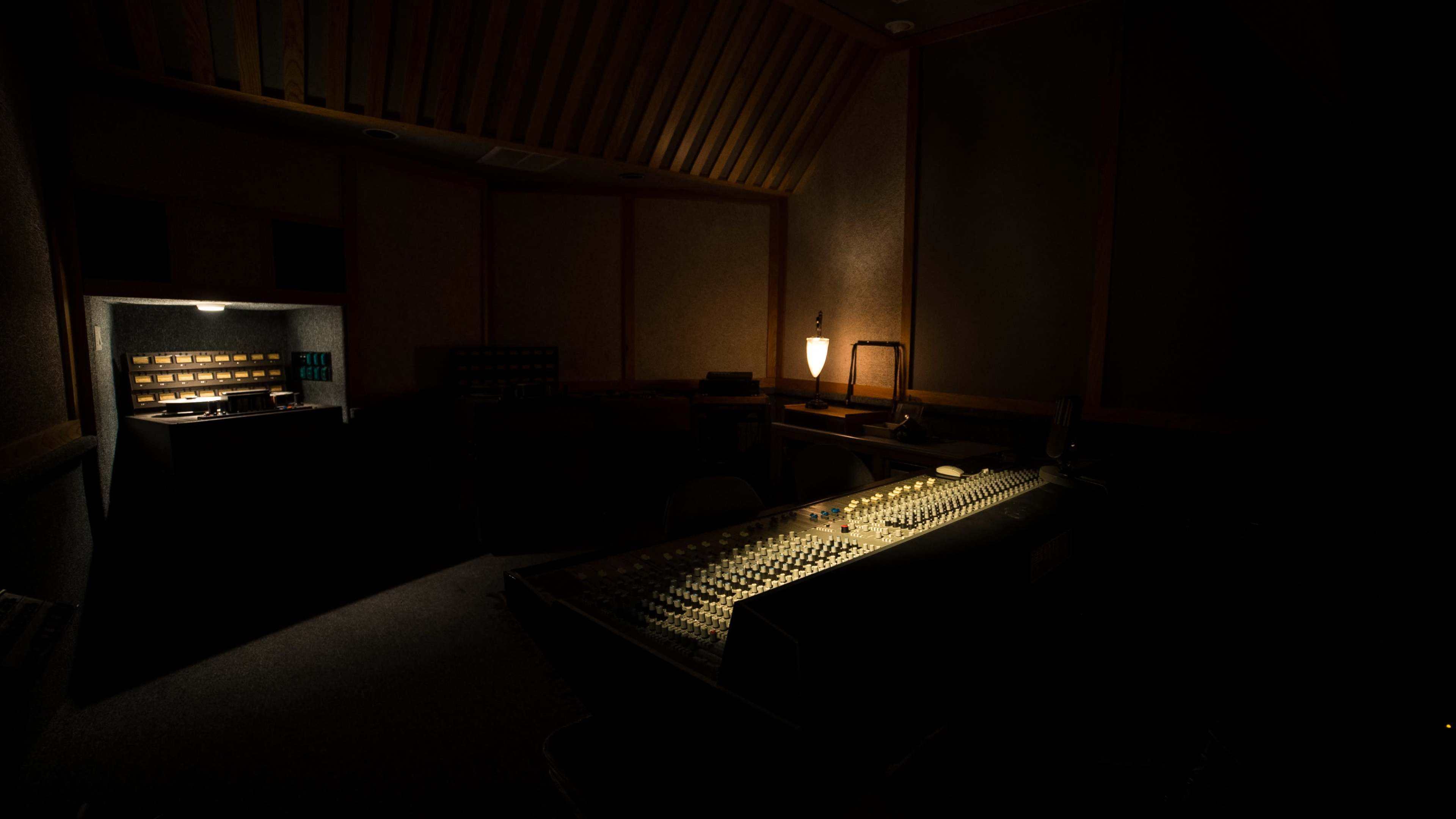 A dimly lit music studio features a large sound mixing console and various audio equipment in shadowy corners.