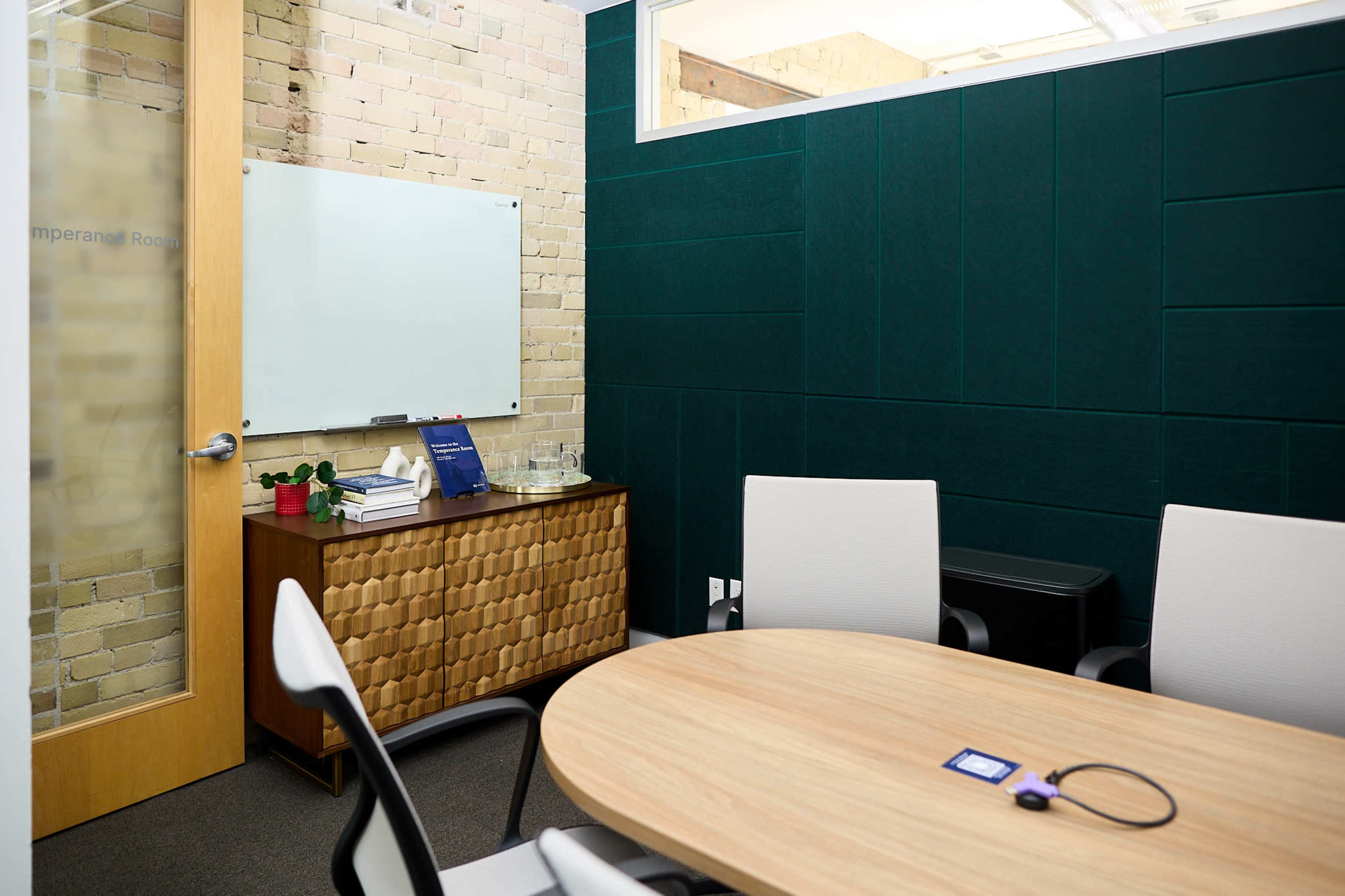 The image depicts a modern conference room with a round wooden table, whiteboard, and textured green wall panels, featuring a glass door and a wooden side cabinet.
