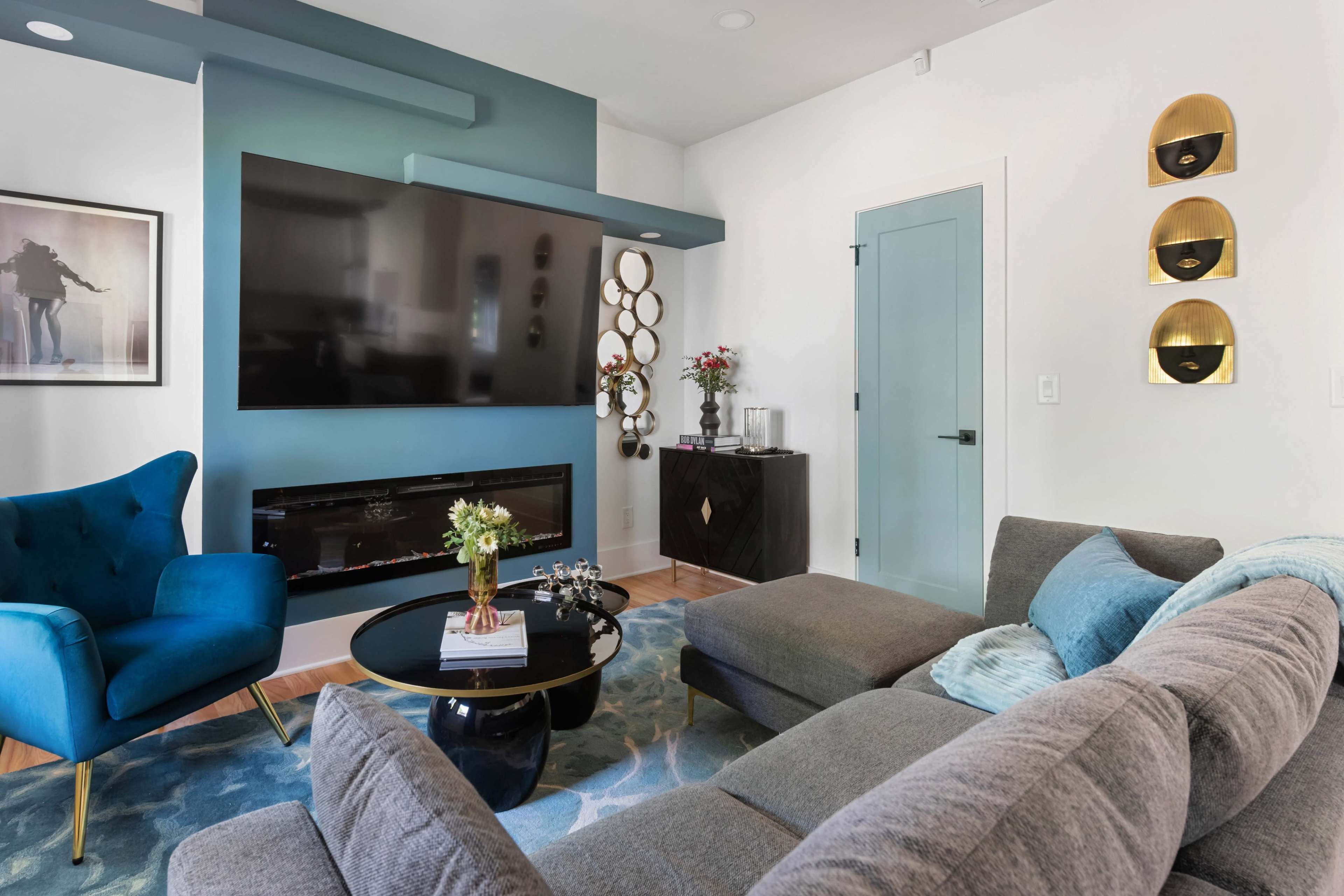 The image shows a modern living room featuring a large flat-screen TV mounted on a blue accent wall, a gray sectional sofa, a round coffee table, and various decorative elements.