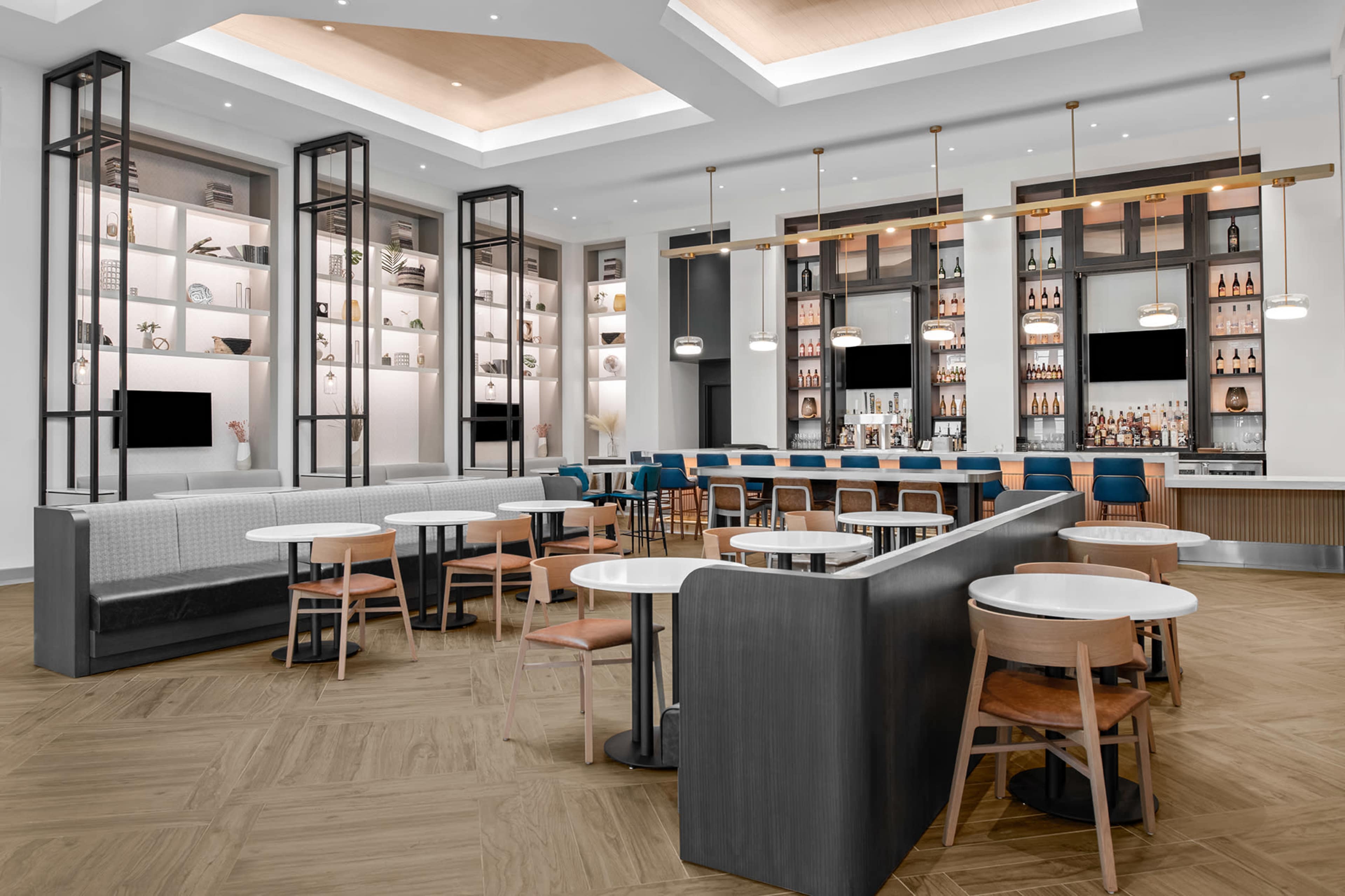 A modern lounge area with a bar, surrounded by booths and tables arranged on a wooden floor under a well-lit ceiling.