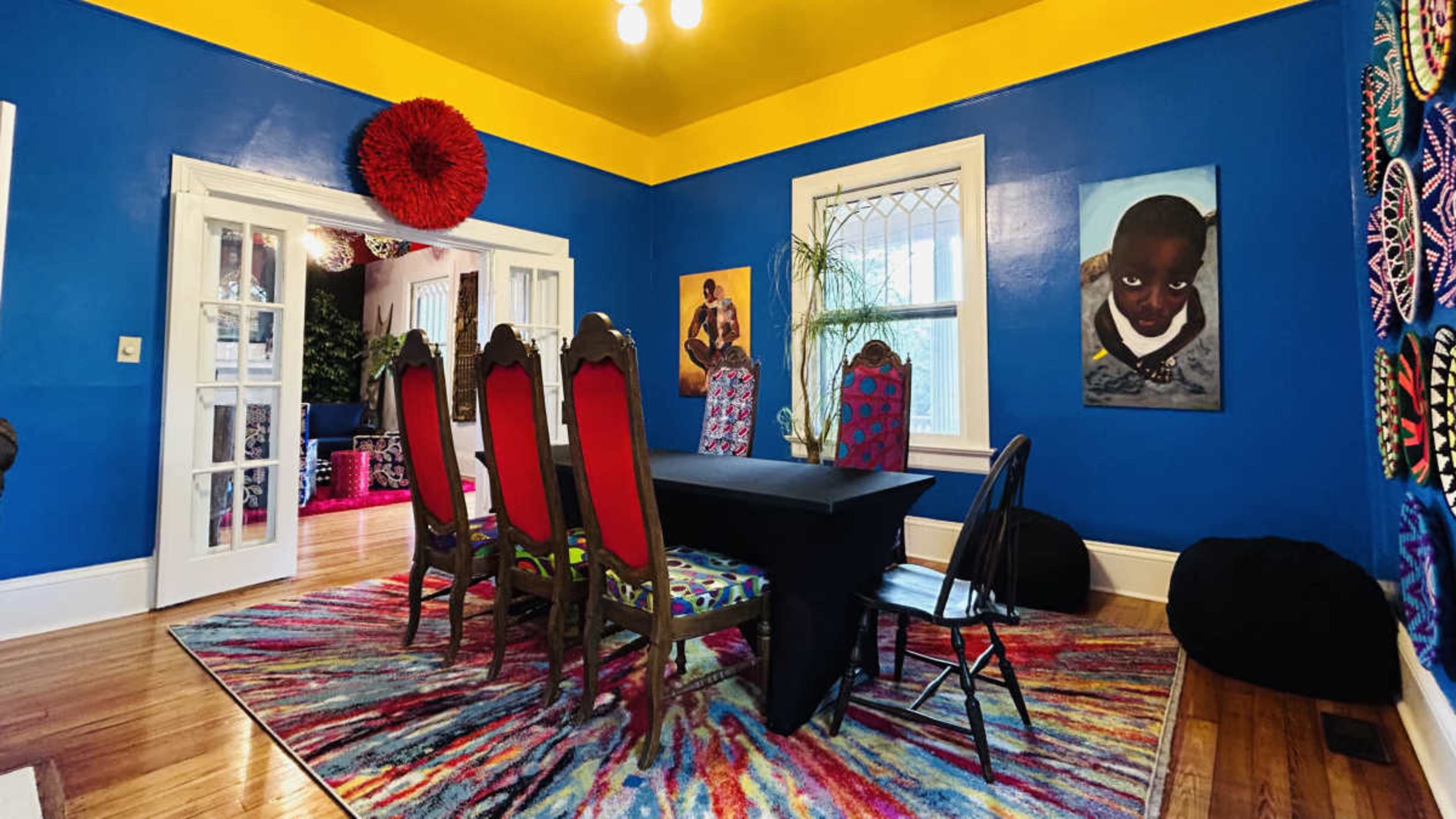 The room features a dining table surrounded by colorful chairs, with vibrant blue walls and a patterned rug.