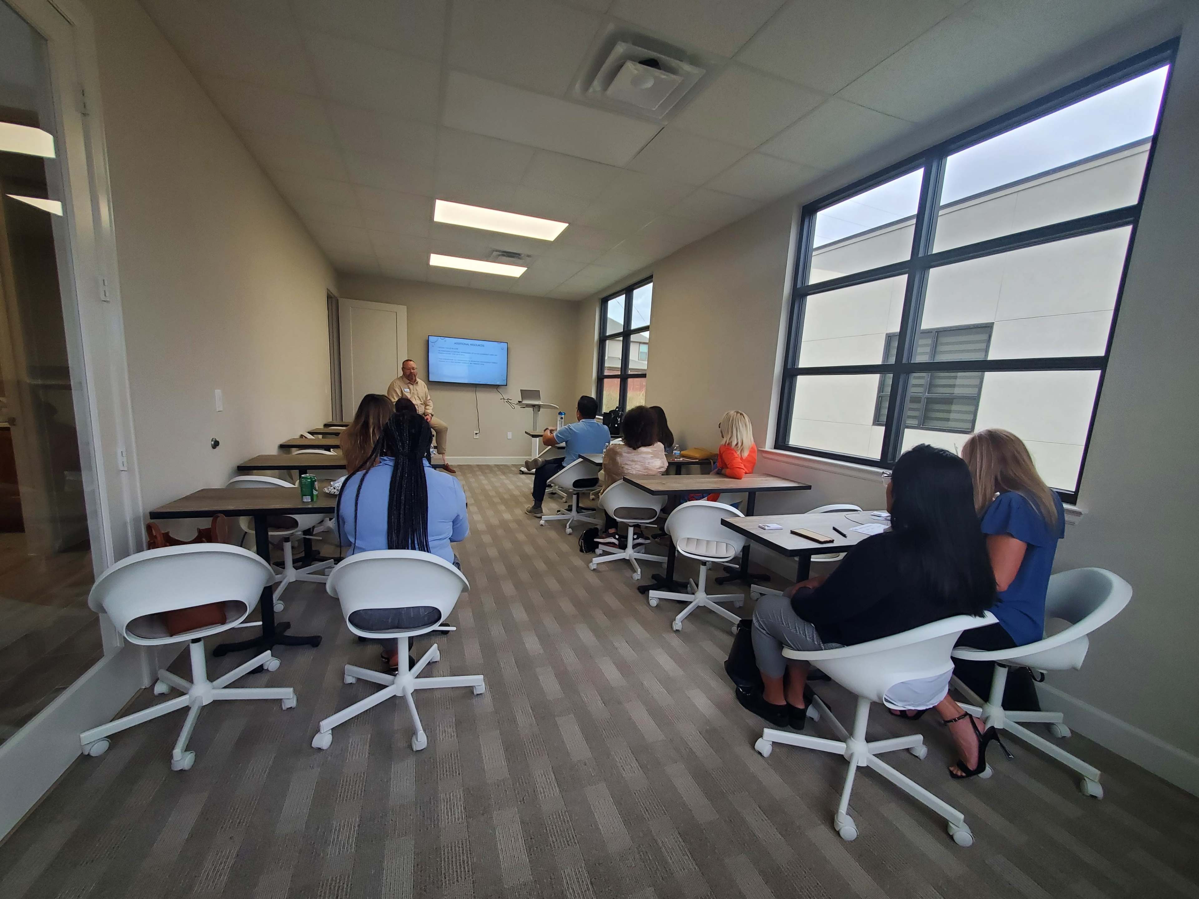 A group of people is seated in a modern classroom, attentively facing a presenter and a large screen displaying information.