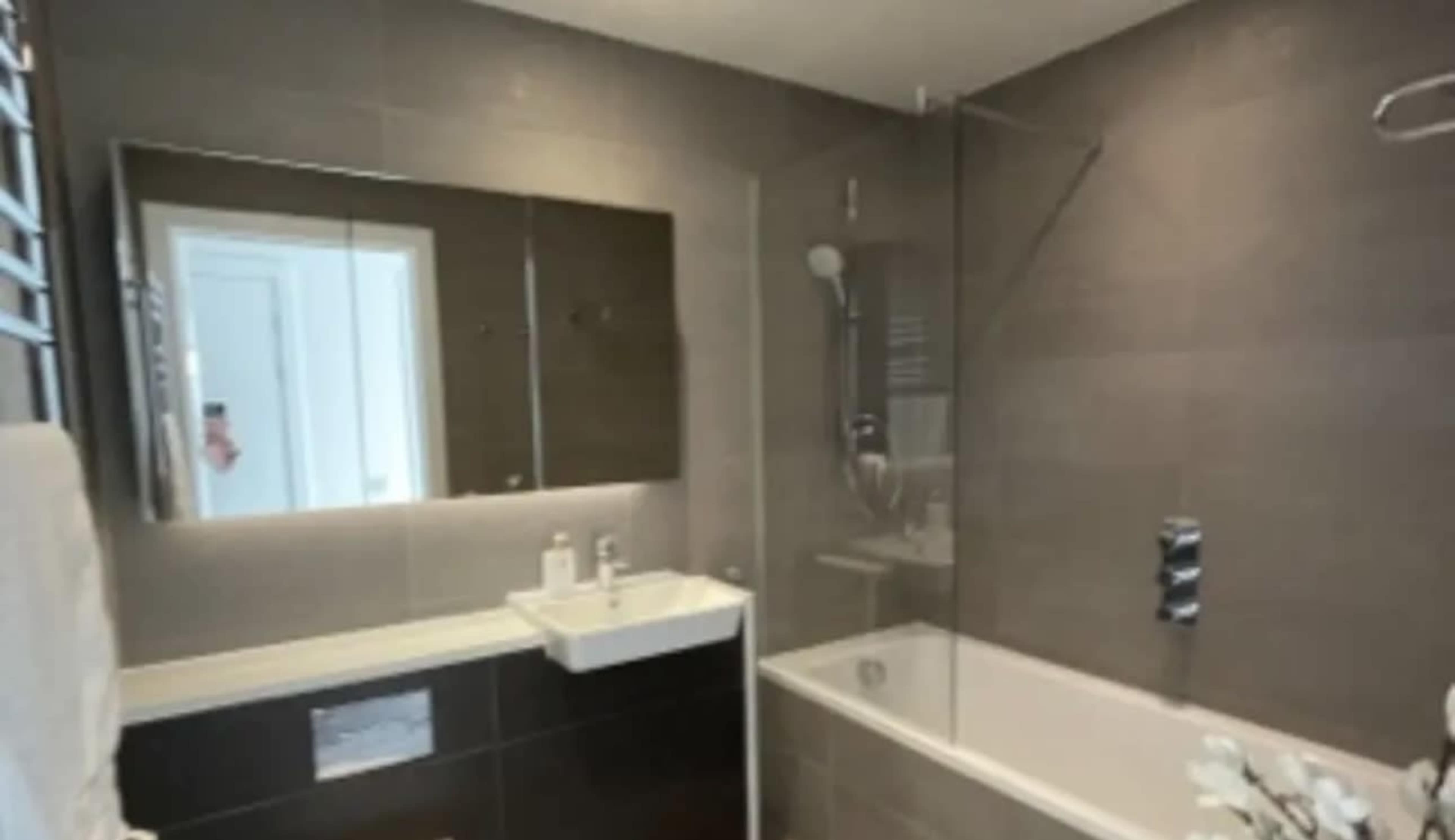 The image shows a modern bathroom featuring a glass shower, a bathtub, a sink with a mirrored cabinet, and dark tiled walls.
