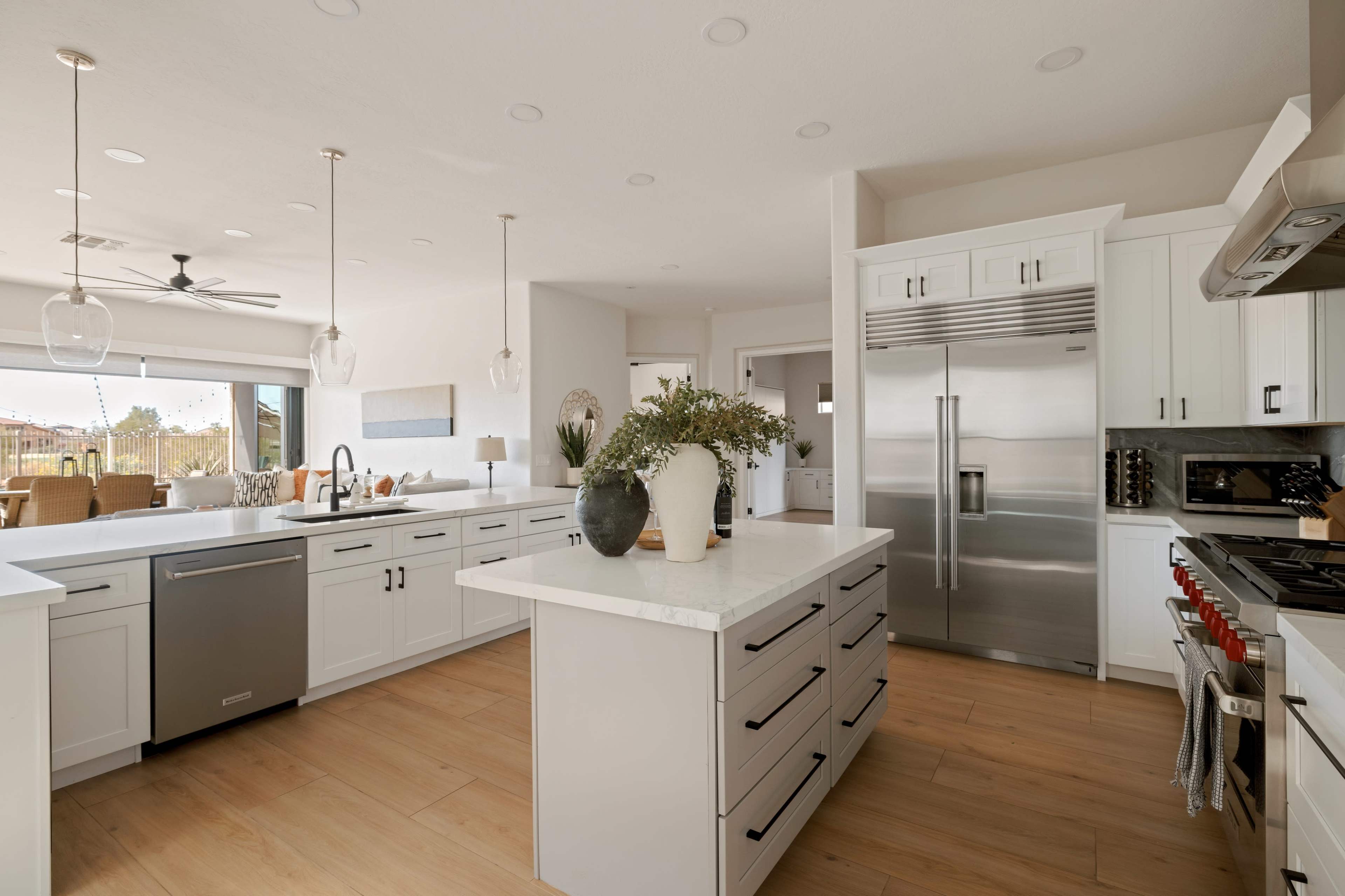 The image shows a modern kitchen featuring an island with a vase of greenery, stainless steel appliances, and white cabinetry, illuminated by pendant lighting.