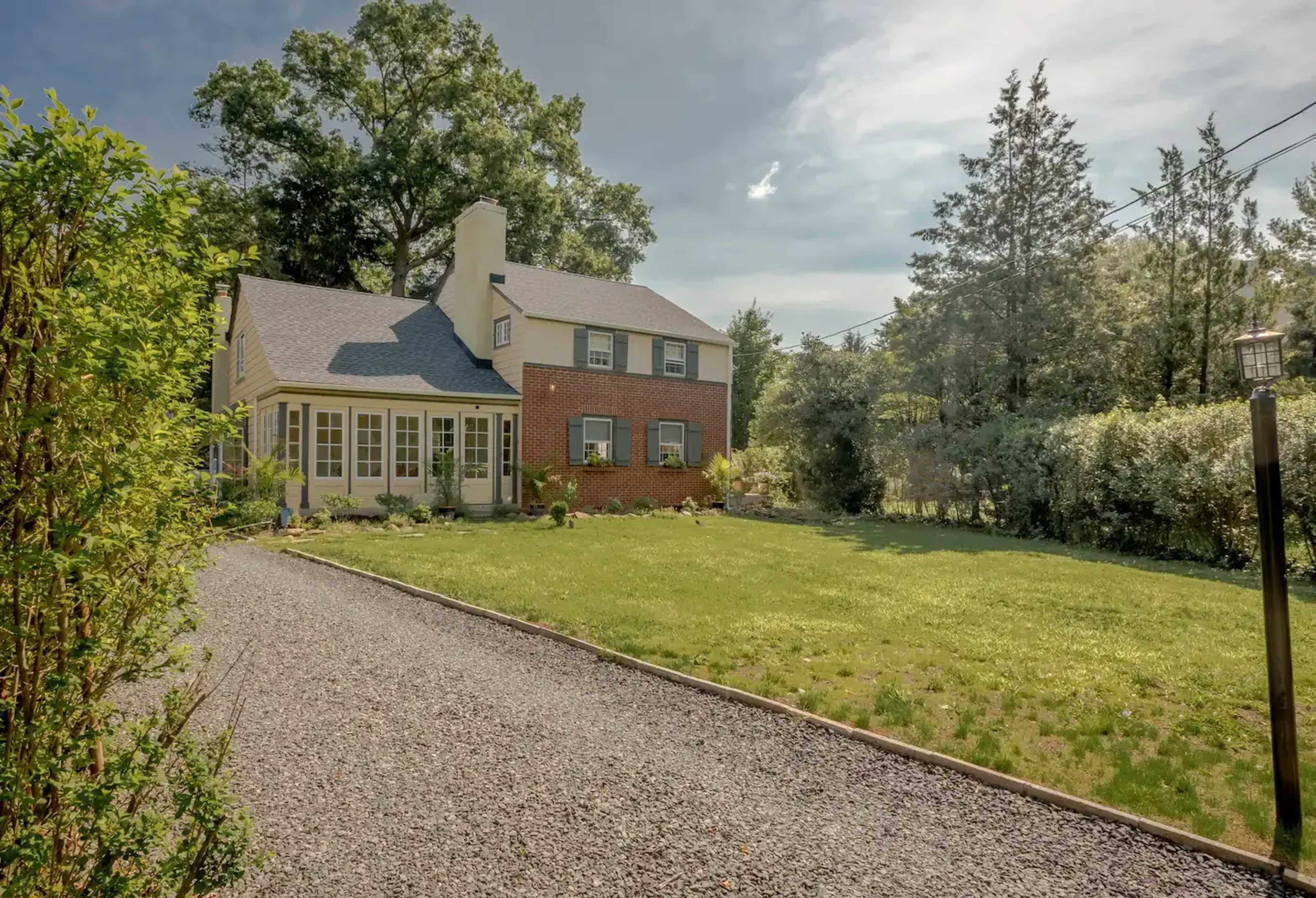 A two-story brick house with a gravel driveway and a green lawn surrounded by trees.
