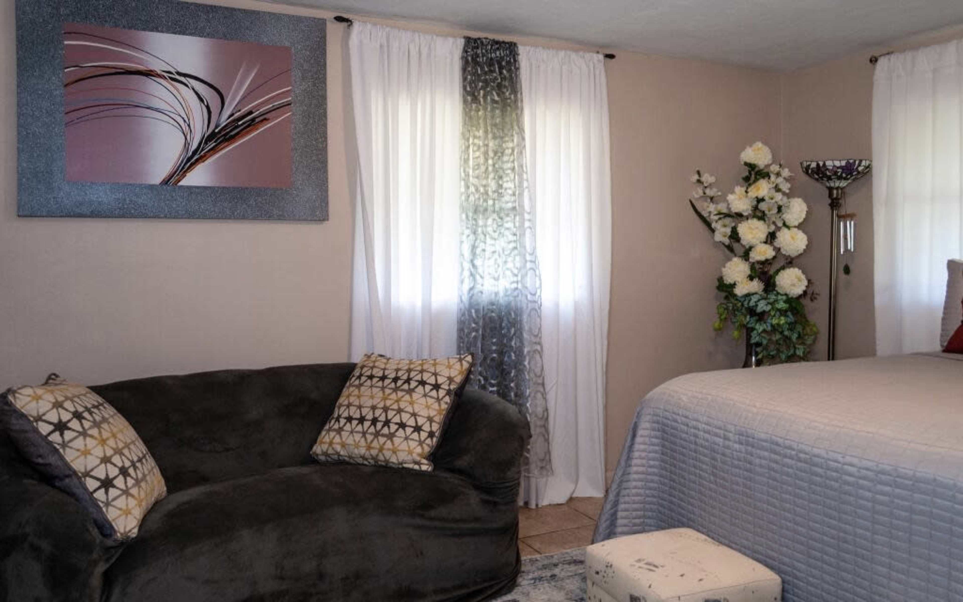 A cozy room with a green sofa, a bed with gray bedding, a floral arrangement, and light-colored curtains.