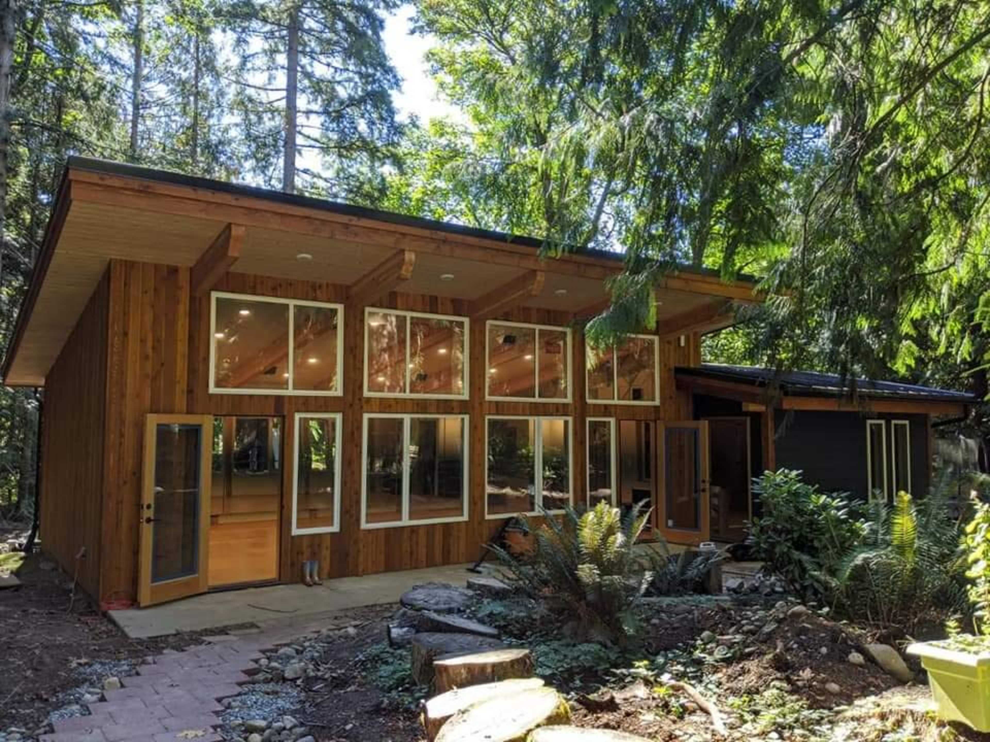 A modern wooden cabin surrounded by trees, featuring large windows and a stone pathway leading to the entrance.