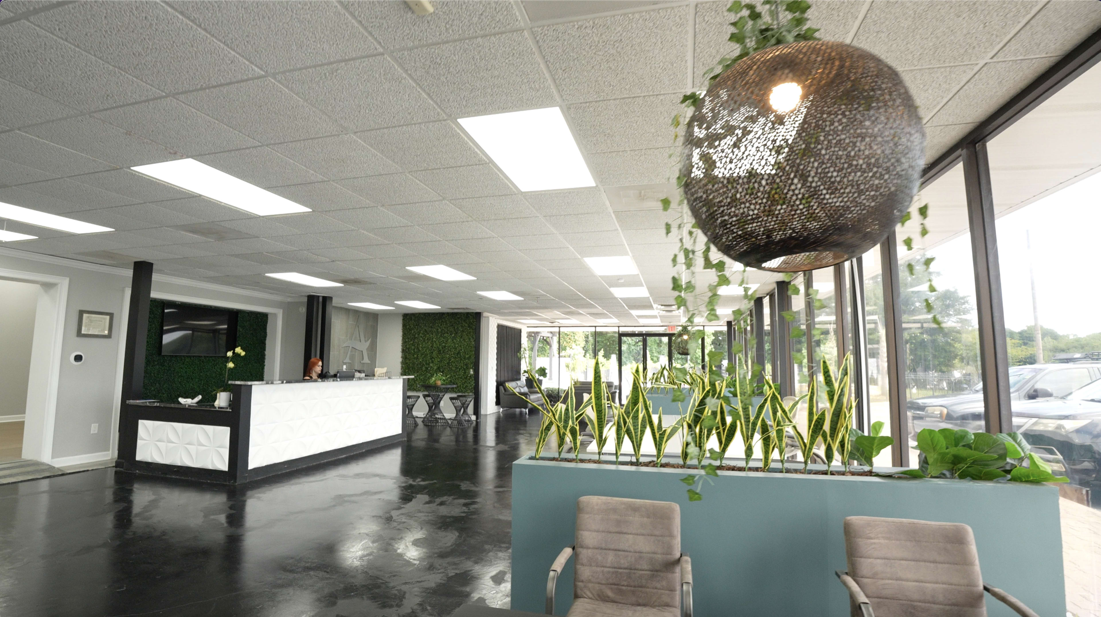 A modern reception area features a geometric front desk, planters with greenery, and large windows allowing natural light.