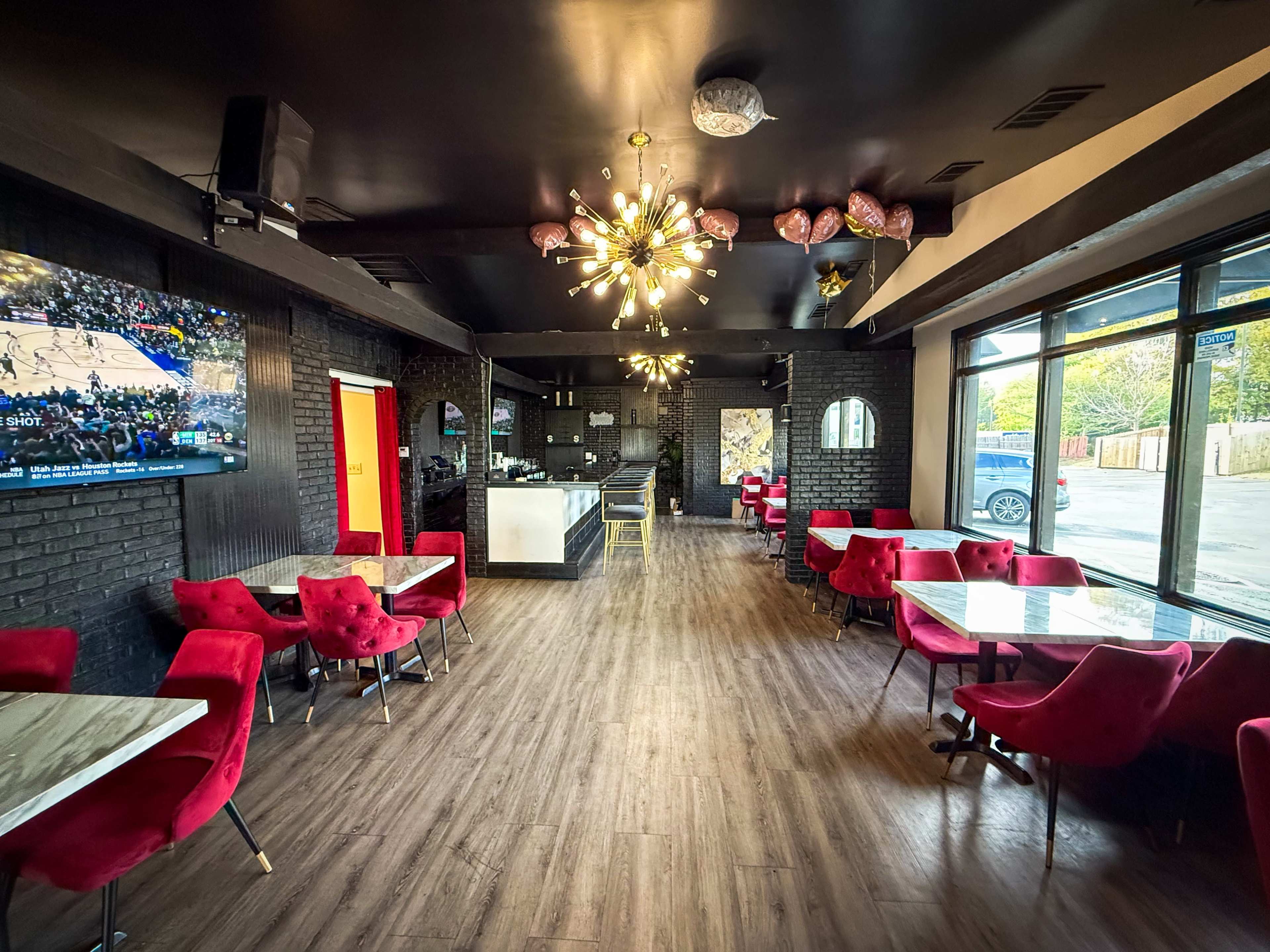 The image shows a modern restaurant interior featuring red chairs, marble-topped tables, and a large television mounted on a black brick wall.