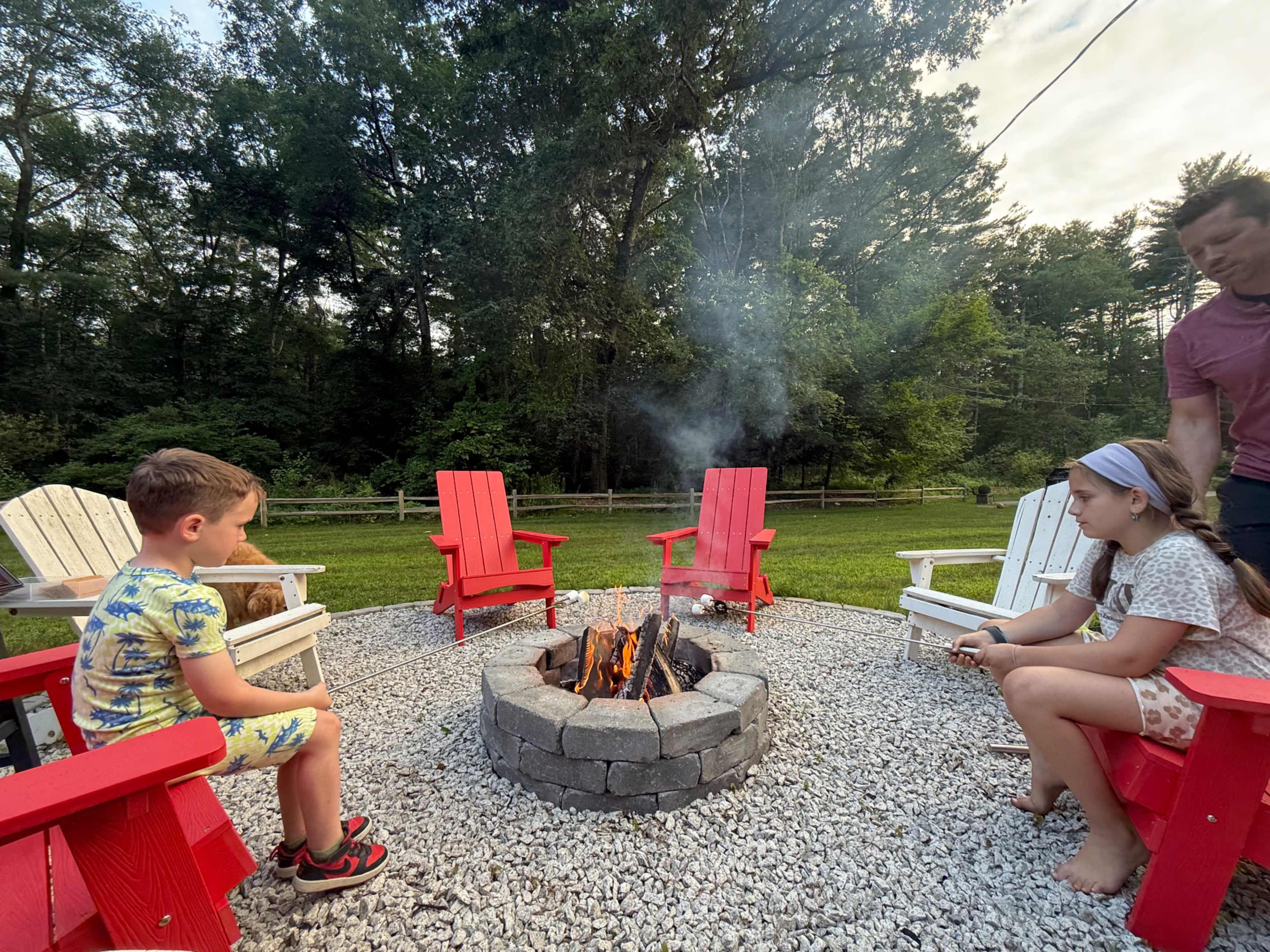 Two children sit on red Adirondack chairs around a stone fire pit with a campfire, while an adult tends to the fire in a backyard setting.