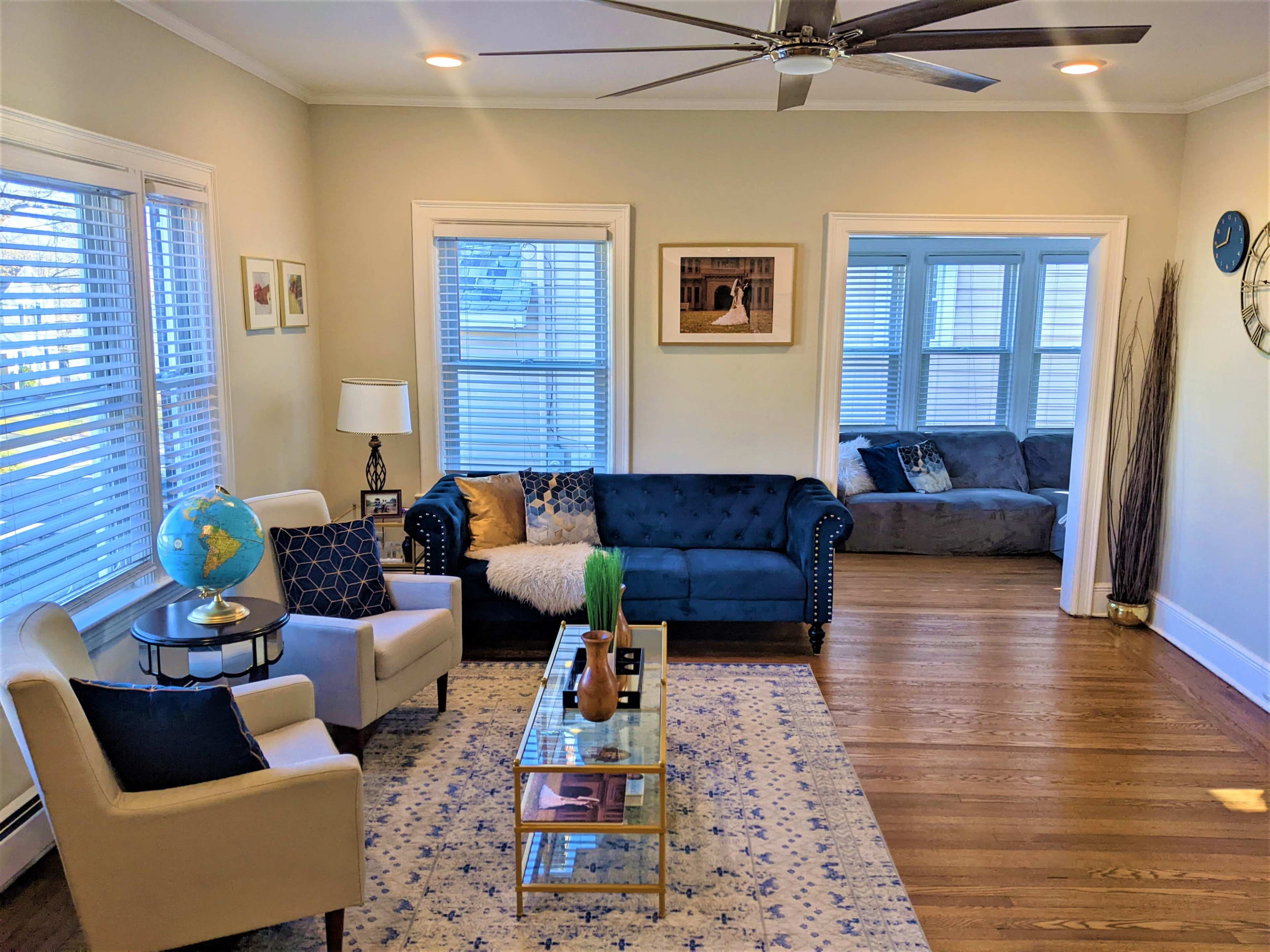 A bright living room featuring a blue tufted sofa, a glass coffee table, two armchairs, and large windows allowing natural light to fill the space.