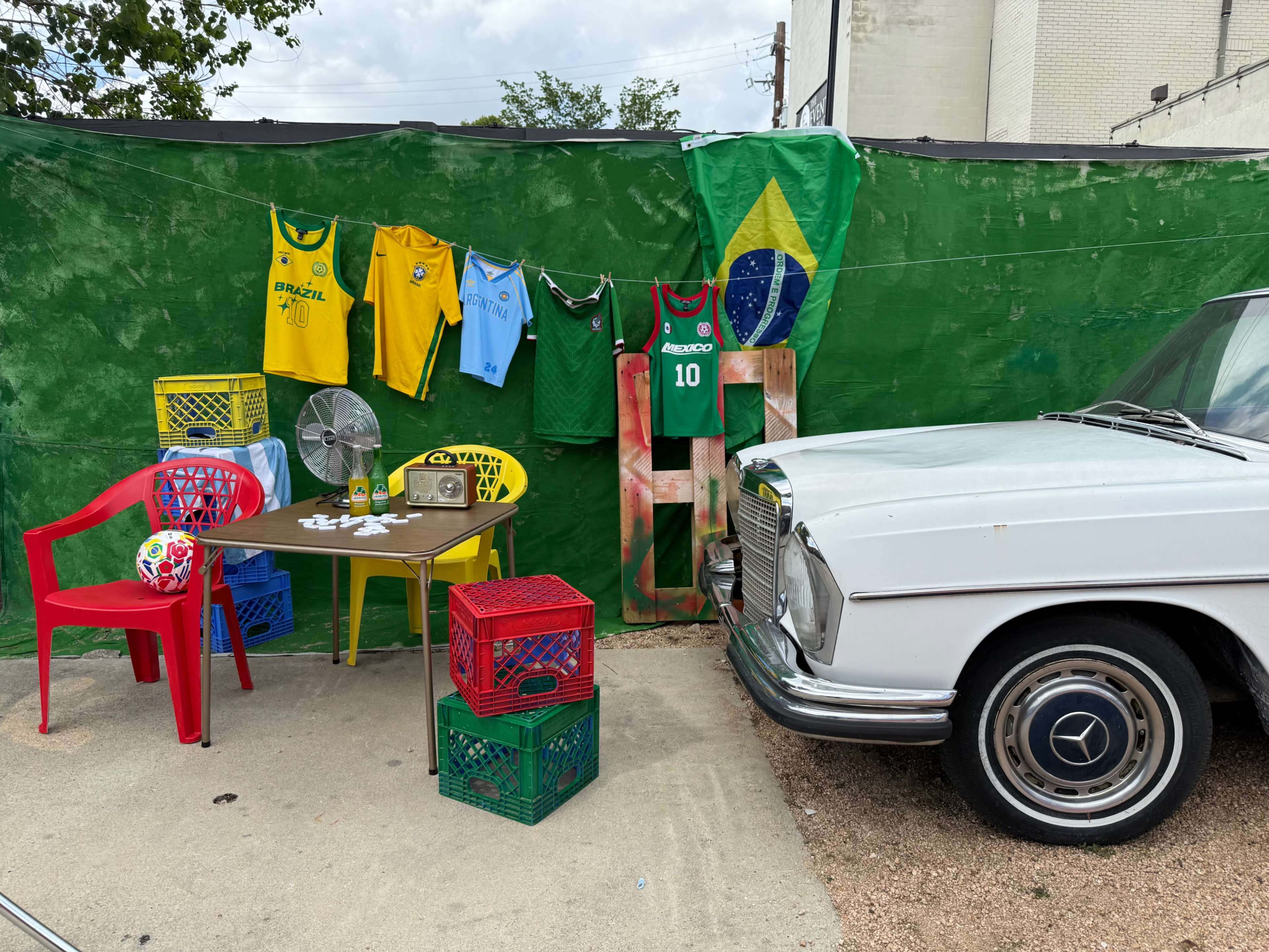 The scene features a vintage white car parked beside colorful plastic chairs and crates arranged under a green tarp, adorned with soccer jerseys and a small fan.