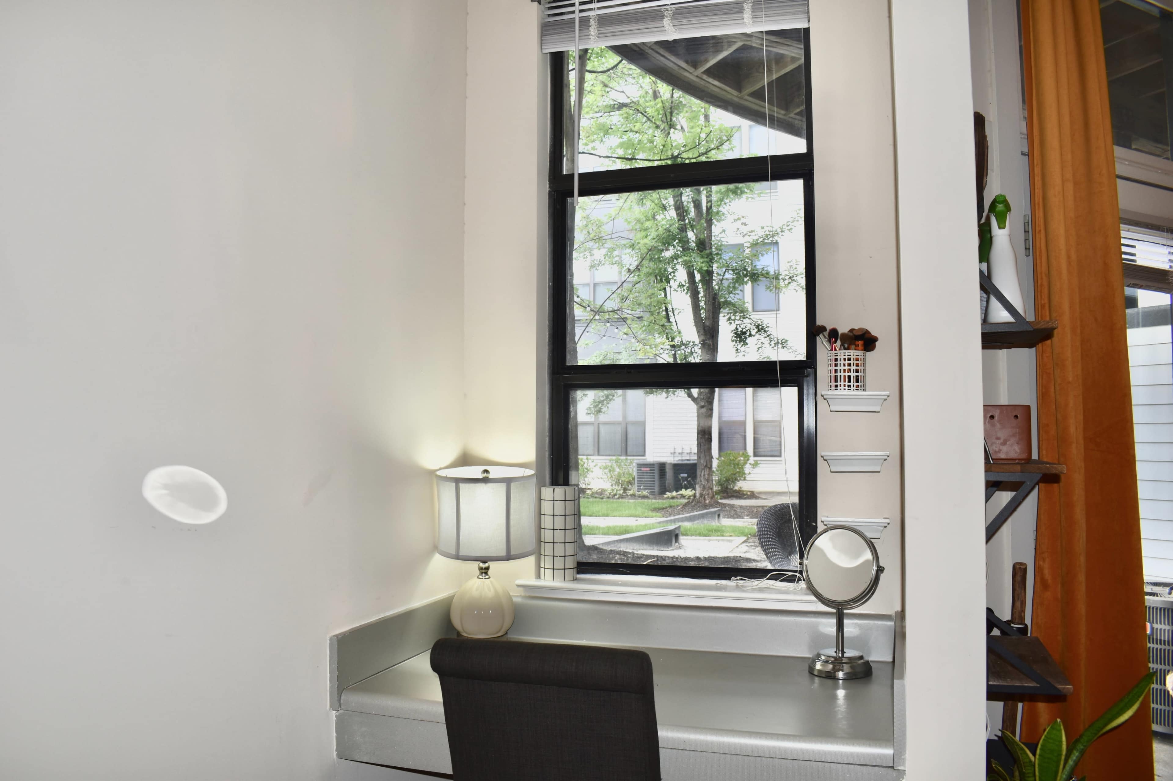 A small workspace with a lamp and mirror is positioned next to a window that overlooks a green outdoor area.