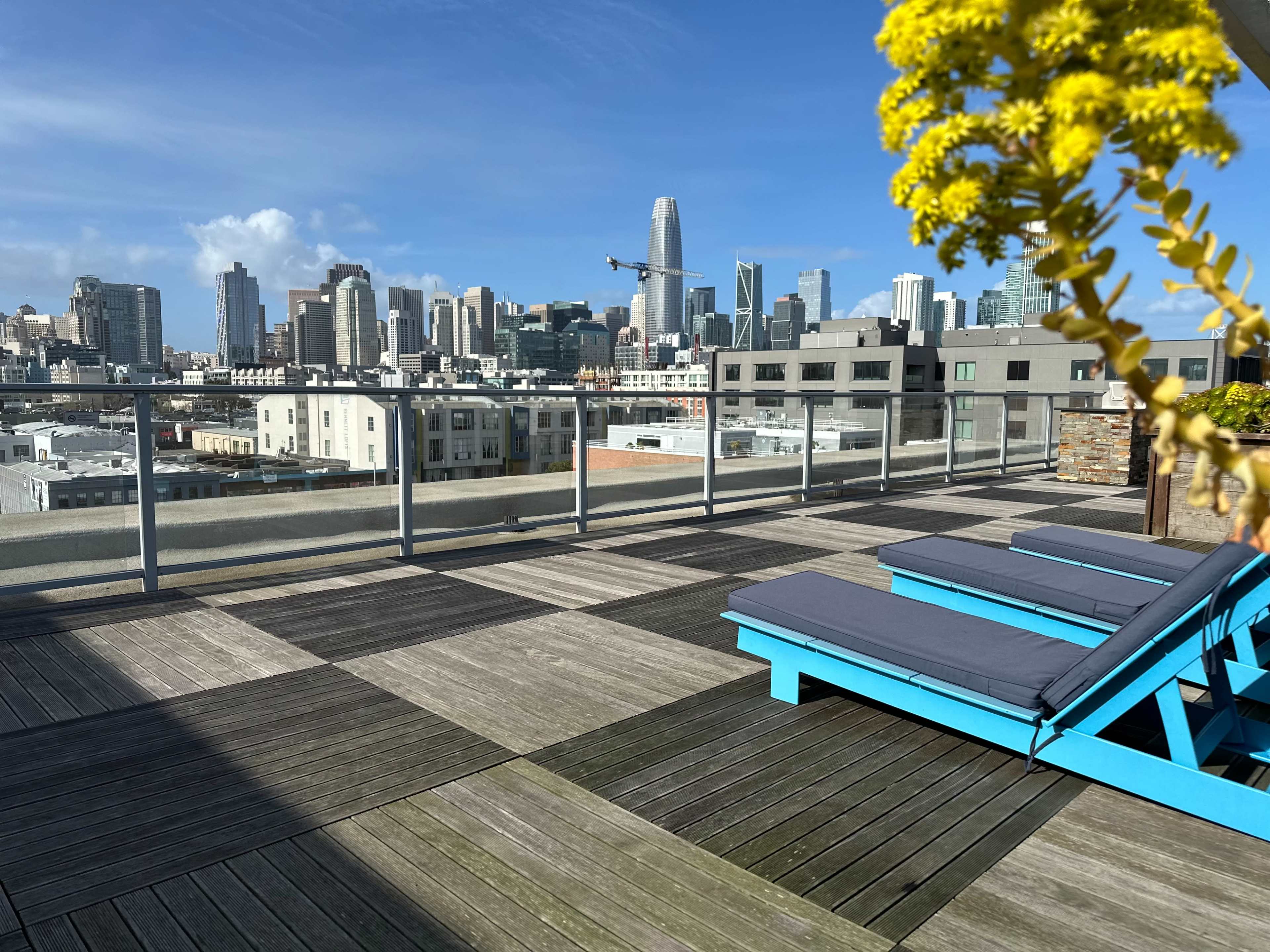 A rooftop terrace with blue lounge chairs overlooks a skyline filled with tall buildings under a clear blue sky.