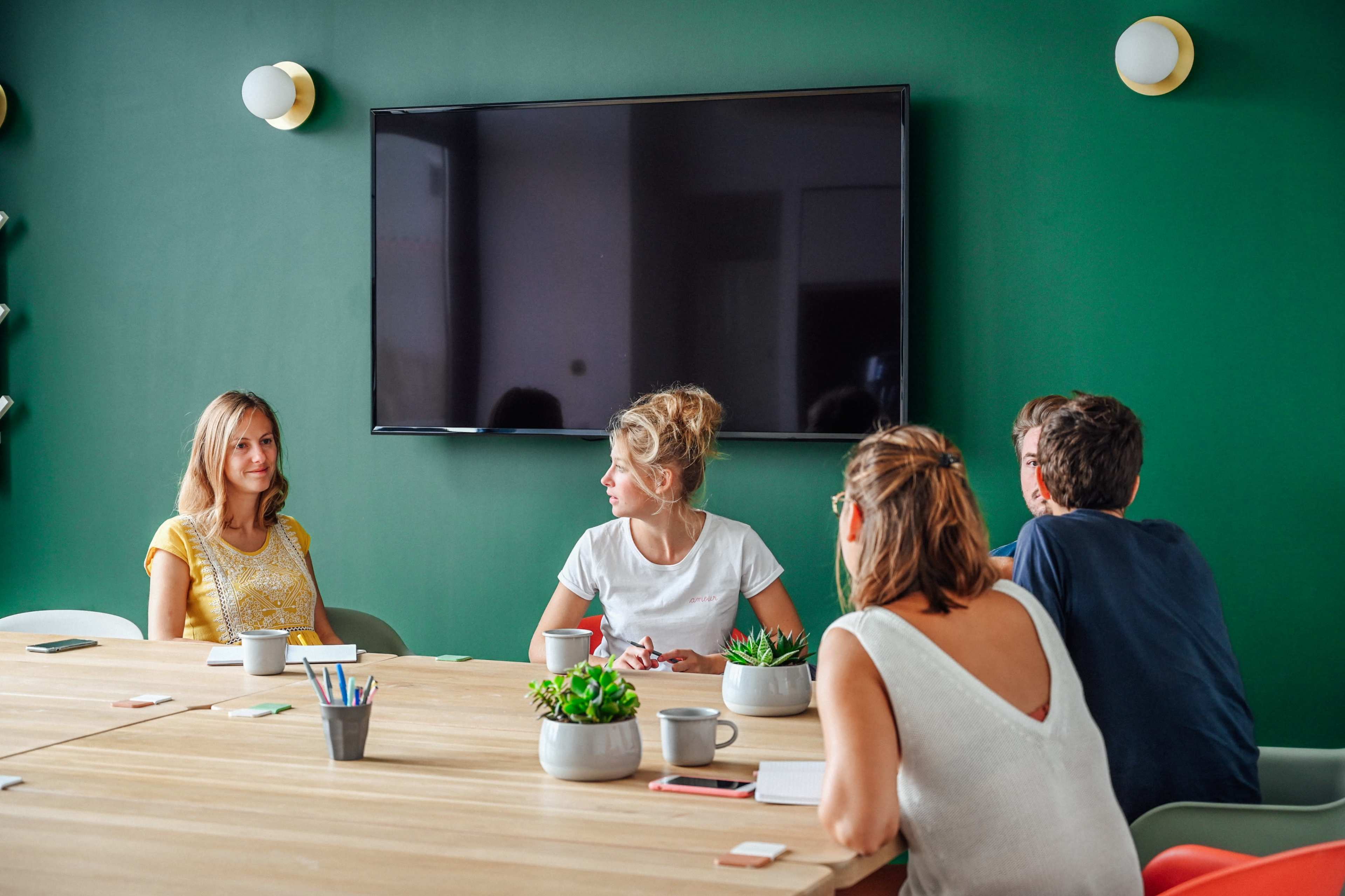 A group of five people sits around a wooden table in a conference room with a green wall and a large screen.