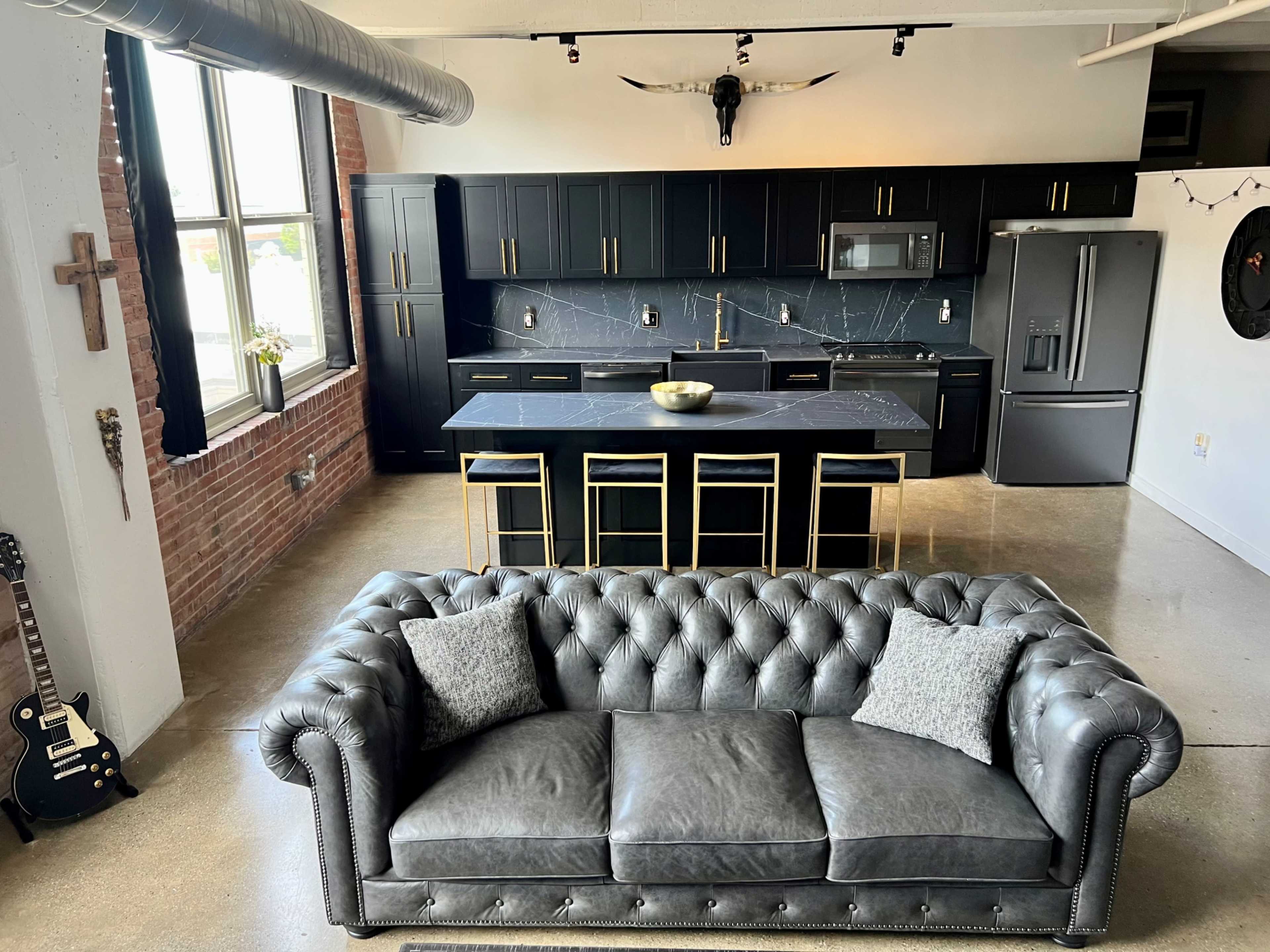 A modern kitchen with black cabinetry and a large island, viewed from above a gray chesterfield sofa.