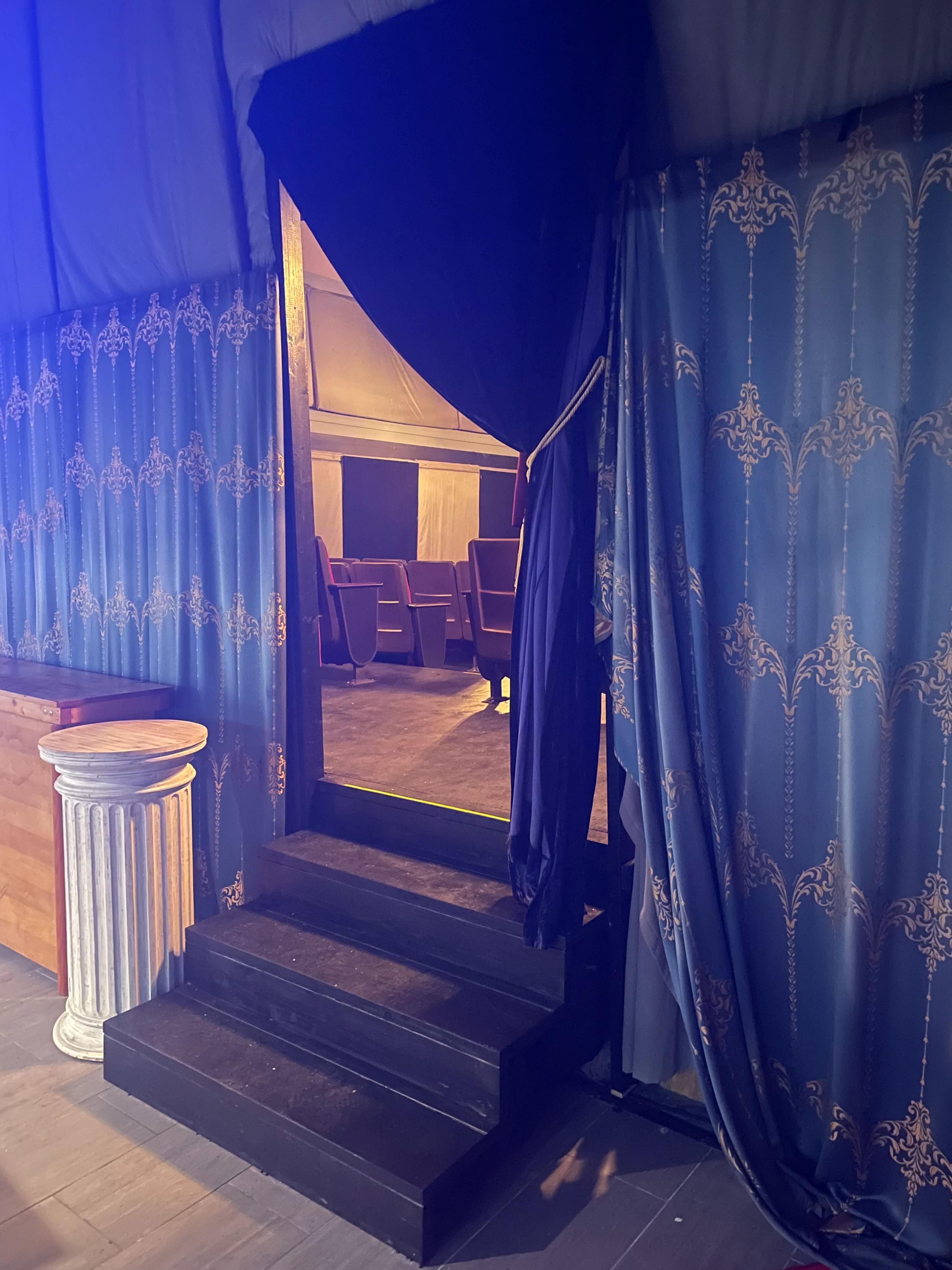 A set of black stairs leads to a doorway draped with blue curtains, revealing a softly lit interior with rows of seating beyond.