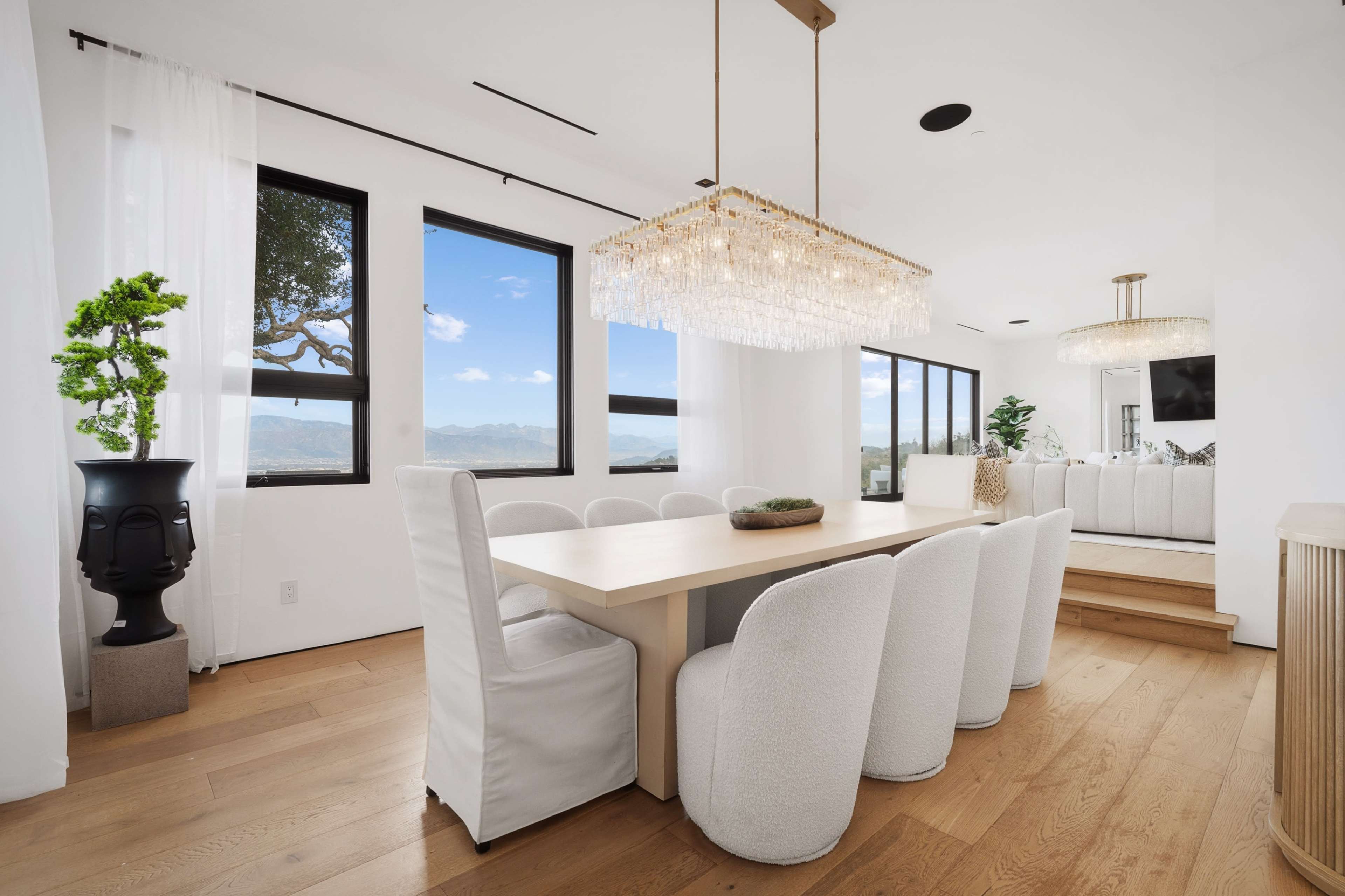 A modern dining area features a long table surrounded by upholstered chairs, with large windows offering a view of the outdoors and two distinct light fixtures above.